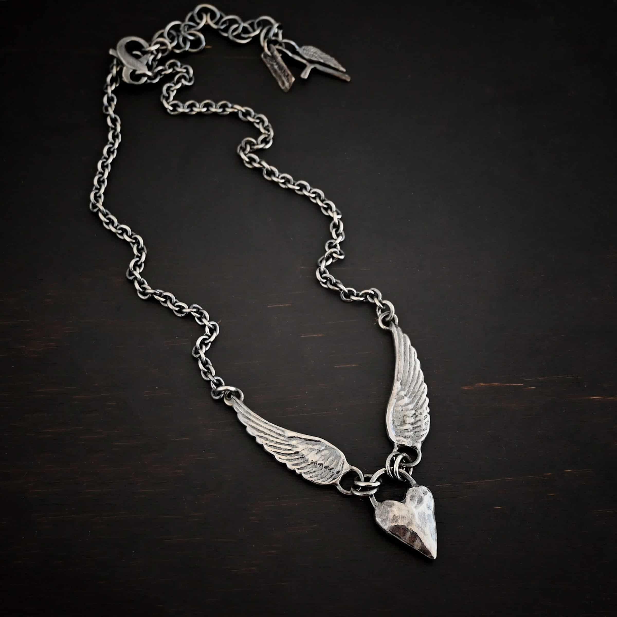 Courage Necklace Photo of the "Courage" Necklace. Made out of sterling silver. There is a thick, solid sterling silver heart that is a medium size for the pendant. It is hammered and oxidized. There is a thick semicircle that is soldered on both sides of the heart on the top. There are four jump rings attached to the semicircle, two on each side. Attached to the jump rings on each side are two angel wings that face opposite directions. The tip of each wing has a loop soldered to the back of them. Attached to the loop on the back of each wing is the chain of the necklace. Necklace is lying against a dark background.