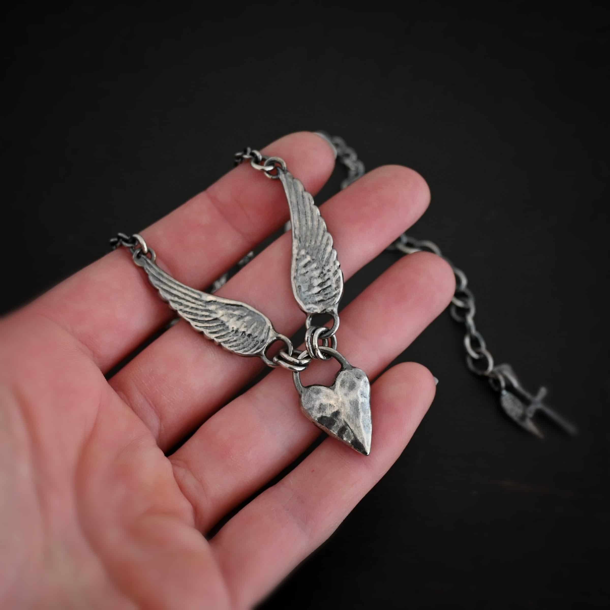 Courage Necklace Photo of the "Courage" Necklace. Made out of sterling silver. There is a thick, solid sterling silver heart that is a medium size for the pendant. It is hammered and oxidized. There is a thick semicircle that is soldered on both sides of the heart on the top. There are four jump rings attached to the semicircle, two on each side. Attached to the jump rings on each side are two angel wings that face opposite directions. The tip of each wing has a loop soldered to the back of them. Attached to the loop on the back of each wing is the chain of the necklace. Necklace is lying against the fingertips of a hand to demonstrate size.