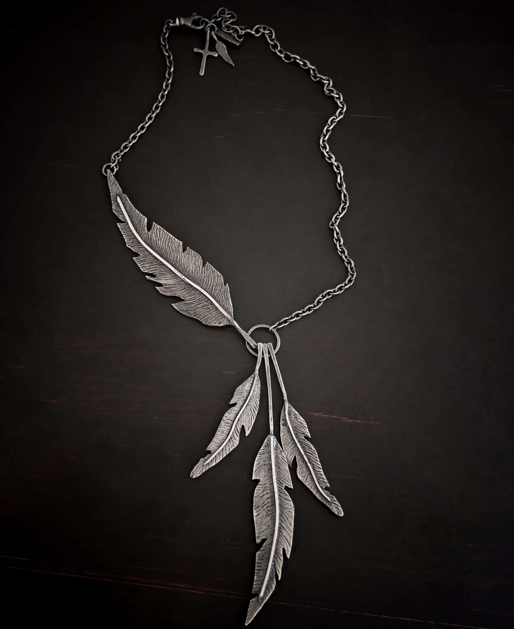 Photo of the "Freedom" Necklace. Made out of sterling silver. There is a medium sized, hammered circle in the center. Hanging from the bottom of it are three large feather charms. Attached to the left side of the circle and hanging horizontally is another large feather charm. It interrupts the chain on the left side for the necklace. The chain attaches to each end of the feather.