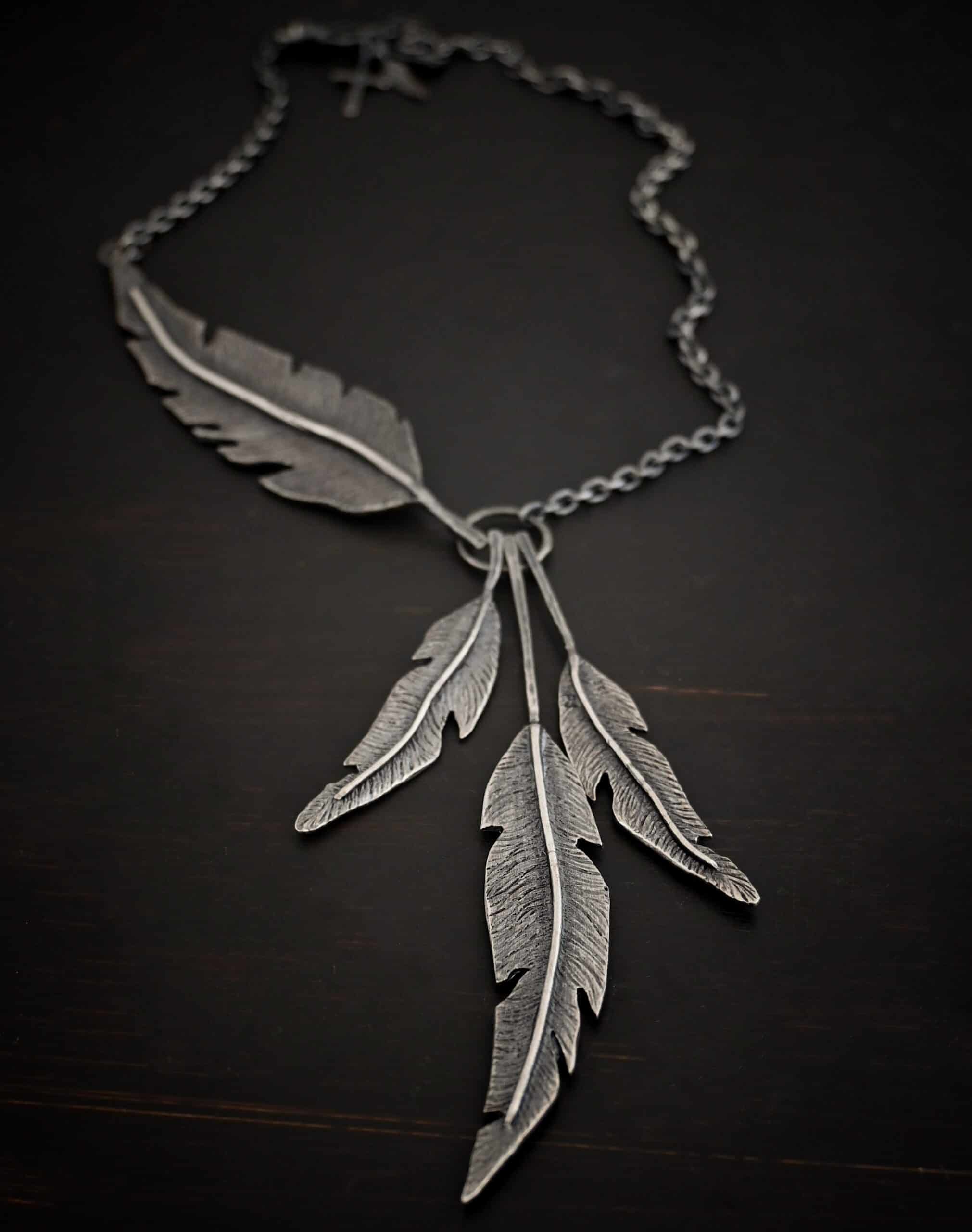 Photo of the "Freedom" Necklace. Made out of sterling silver. There is a medium sized, hammered circle in the center. Hanging from the bottom of it are three large feather charms. Attached to the left side of the circle and hanging horizontally is another large feather charm. It interrupts the chain on the left side for the necklace. The chain attaches to each end of the feather.
