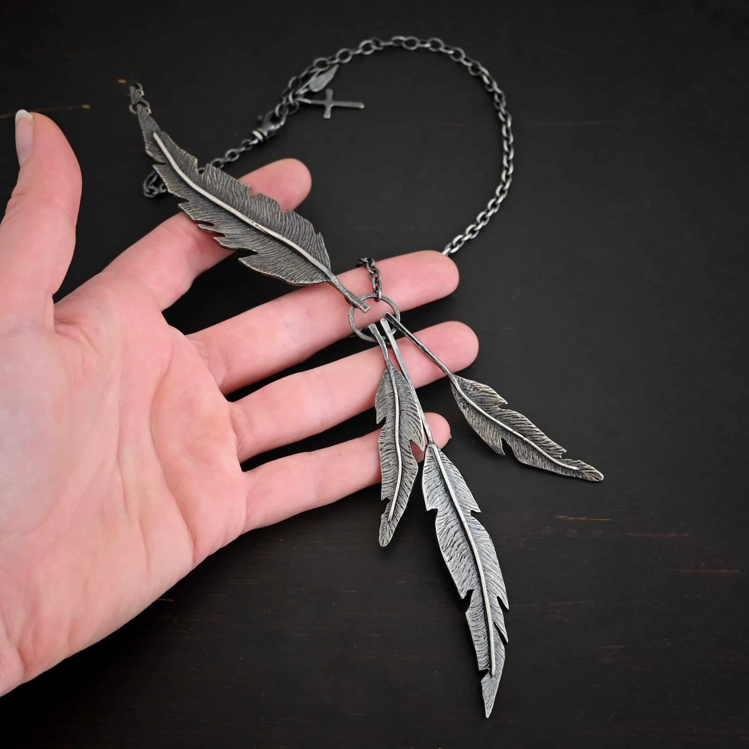 Photo of the "Freedom" Necklace. Made out of sterling silver. There is a medium sized, hammered circle in the center. Hanging from the bottom of it are three large feather charms. Attached to the left side of the circle and hanging horizontally is another large feather charm. It interrupts the chain on the left side for the necklace. The chain attaches to each end of the feather. Necklace is lying against the fingertips of a hand.
