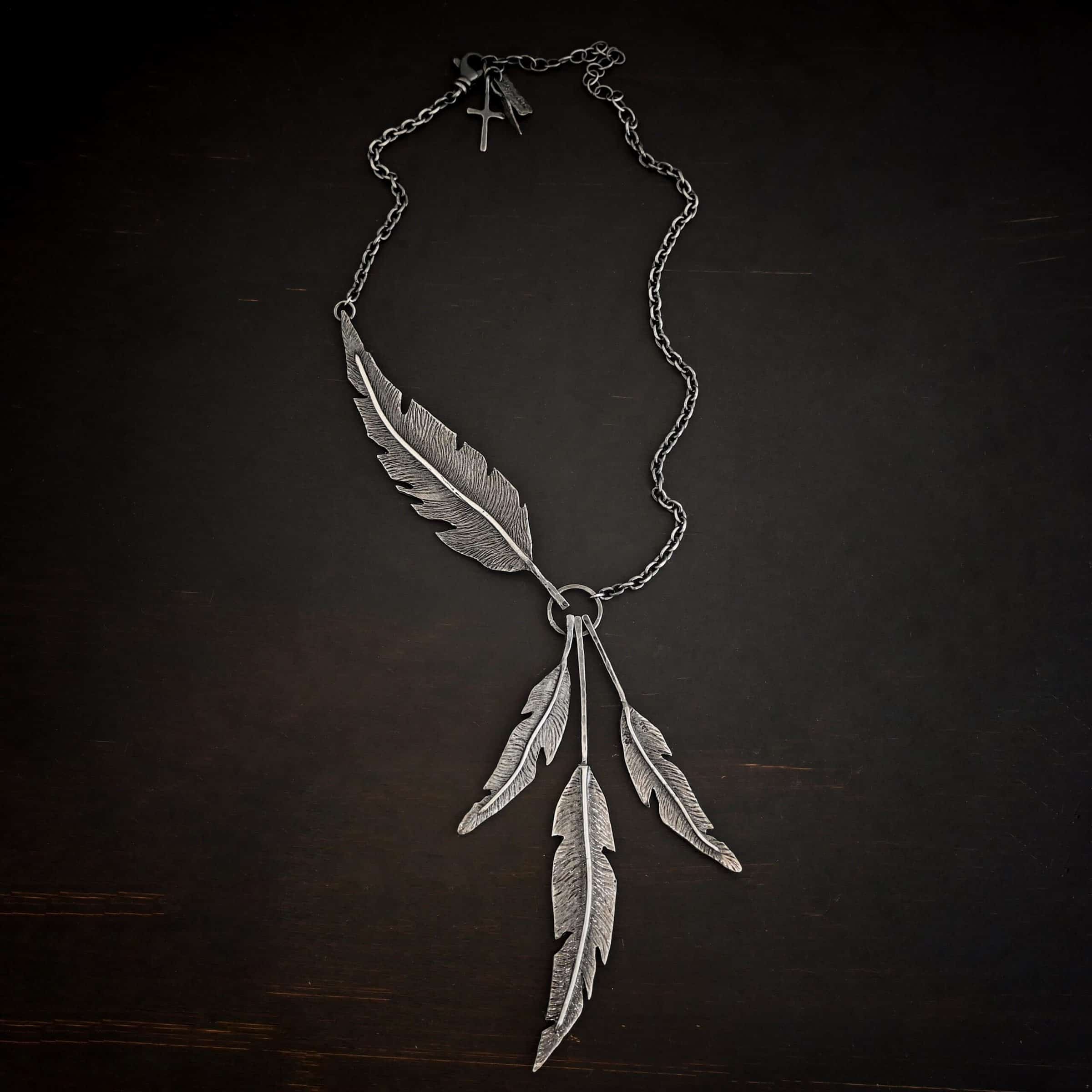 Photo of the "Freedom" Necklace. Made out of sterling silver. There is a medium sized, hammered circle in the center. Hanging from the bottom of it are three large feather charms. Attached to the left side of the circle and hanging horizontally is another large feather charm. It interrupts the chain on the left side for the necklace. The chain attaches to each end of the feather.
