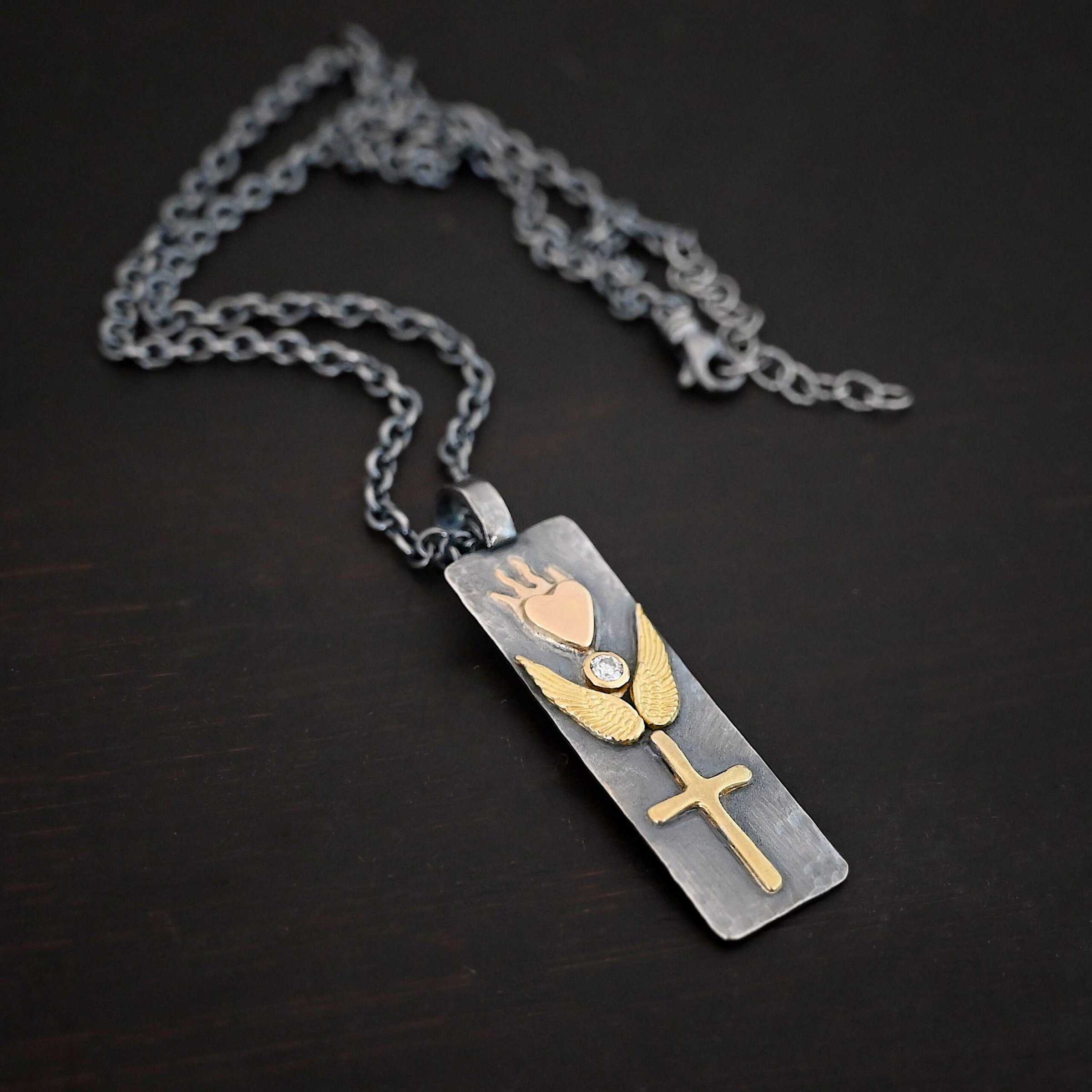 Protect Necklace Photo of the "Protect" Necklace. Necklace is made out of silver and gold. The pendant is a rectangle shaped silver tag that hangs vertically. It is large. On the top of the rectangle is a gold heart with gold flames dancing across the top of it. On the bottom, beneath the heart is a diamond that is set in gold. On each side of the diamond is a gold angel wing. Beneath the gold angel wings in the center of them and on the bottom of the rectangle pendant is a gold cross. There is a thick jump ring soldered to the top of the pendant that the chain of the necklace goes through. Necklace is laying against a dark background.
