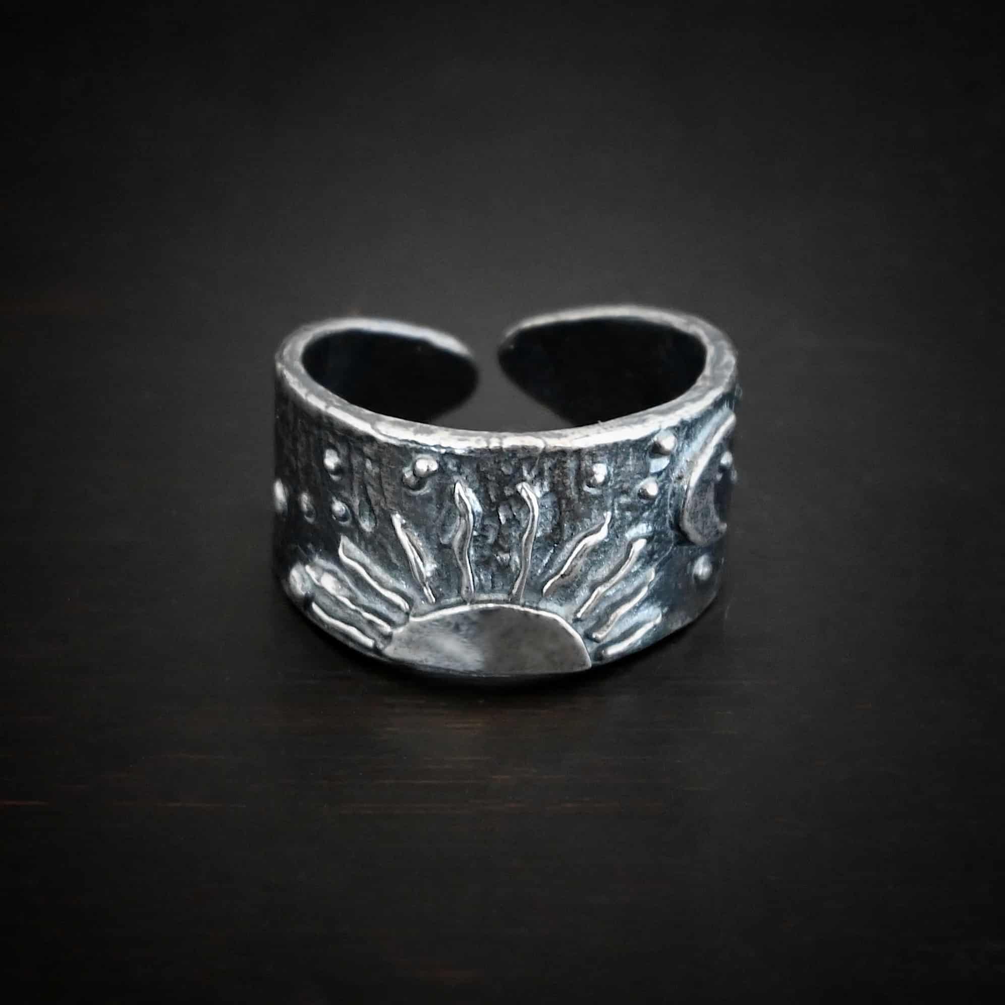 Sun Moon Stars Ring Photo of the "Sun Moon Stars" Ring. Made out of sterling silver. The bottom of the ring in the center has the top half of a rising sun. There are ten short silver lines that are wavy that go around this to signify the sun rising. There is a crescent moon on the top right side of the band. All around the band, there is silver ball detail. Ring is sitting against a dark background.