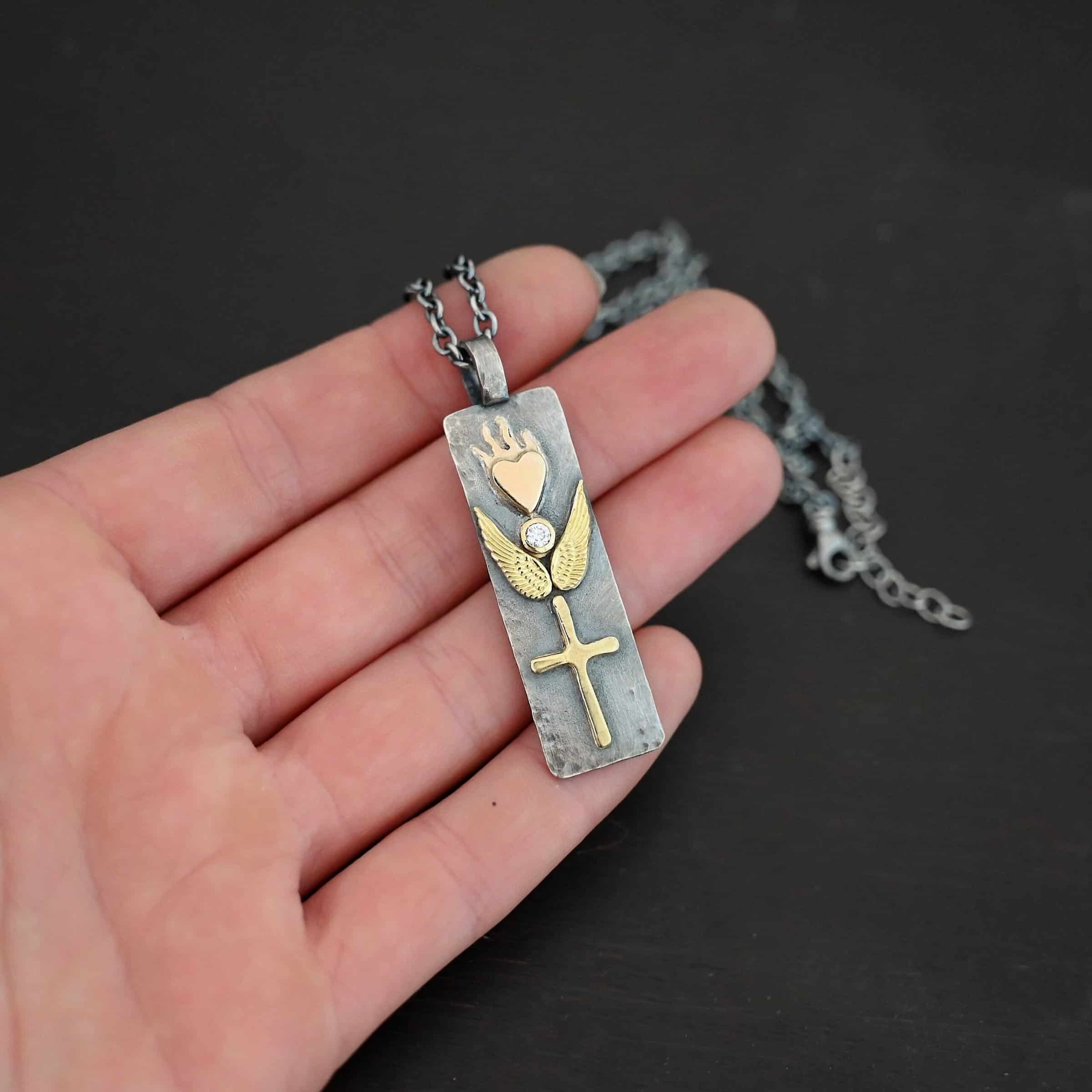 Protect Necklace Photo of the "Protect" Necklace. Necklace is made out of silver and gold. The pendant is a rectangle shaped silver tag that hangs vertically. It is large. On the top of the rectangle is a gold heart with gold flames dancing across the top of it. On the bottom, beneath the heart is a diamond that is set in gold. On each side of the diamond is a gold angel wing. Beneath the gold angel wings in the center of them and on the bottom of the rectangle pendant is a gold cross. There is a thick jump ring soldered to the top of the pendant that the chain of the necklace goes through. Necklace is laying against the fingertips of a hand to demonstrate size.