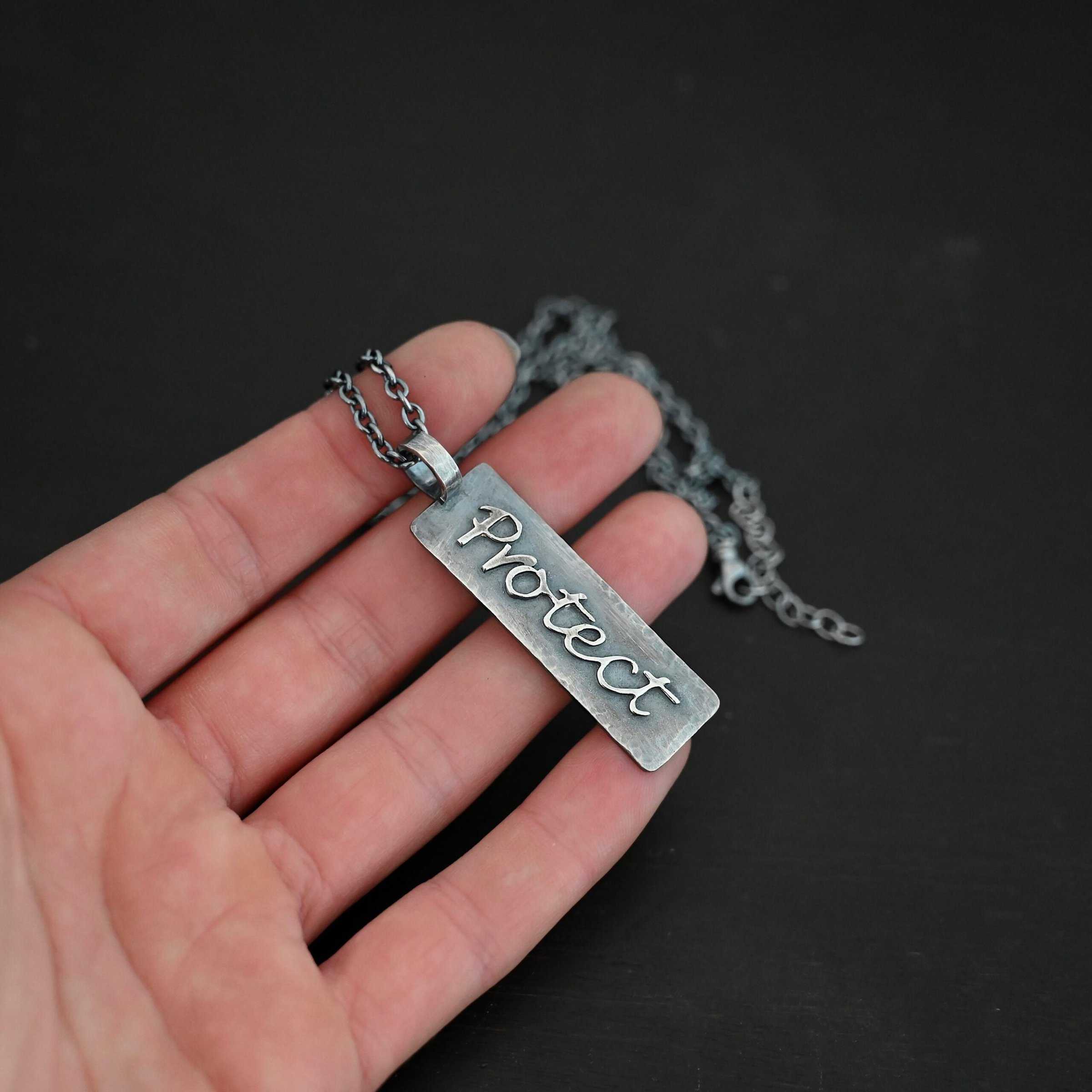 Protect Necklace Photo of the back of the "Protect" Necklace. Necklace is made out of sterling silver. The pendant is a rectangle shaped silver tag that hangs vertically. It is large. The word "Protect" is written across the entire rectangle in carved out silver and soldered to the rectangle. There is a thick jump ring soldered to the top of the pendant that the chain of the necklace goes through. Necklace is laying against the fingertips of a hand to demonstrate size.