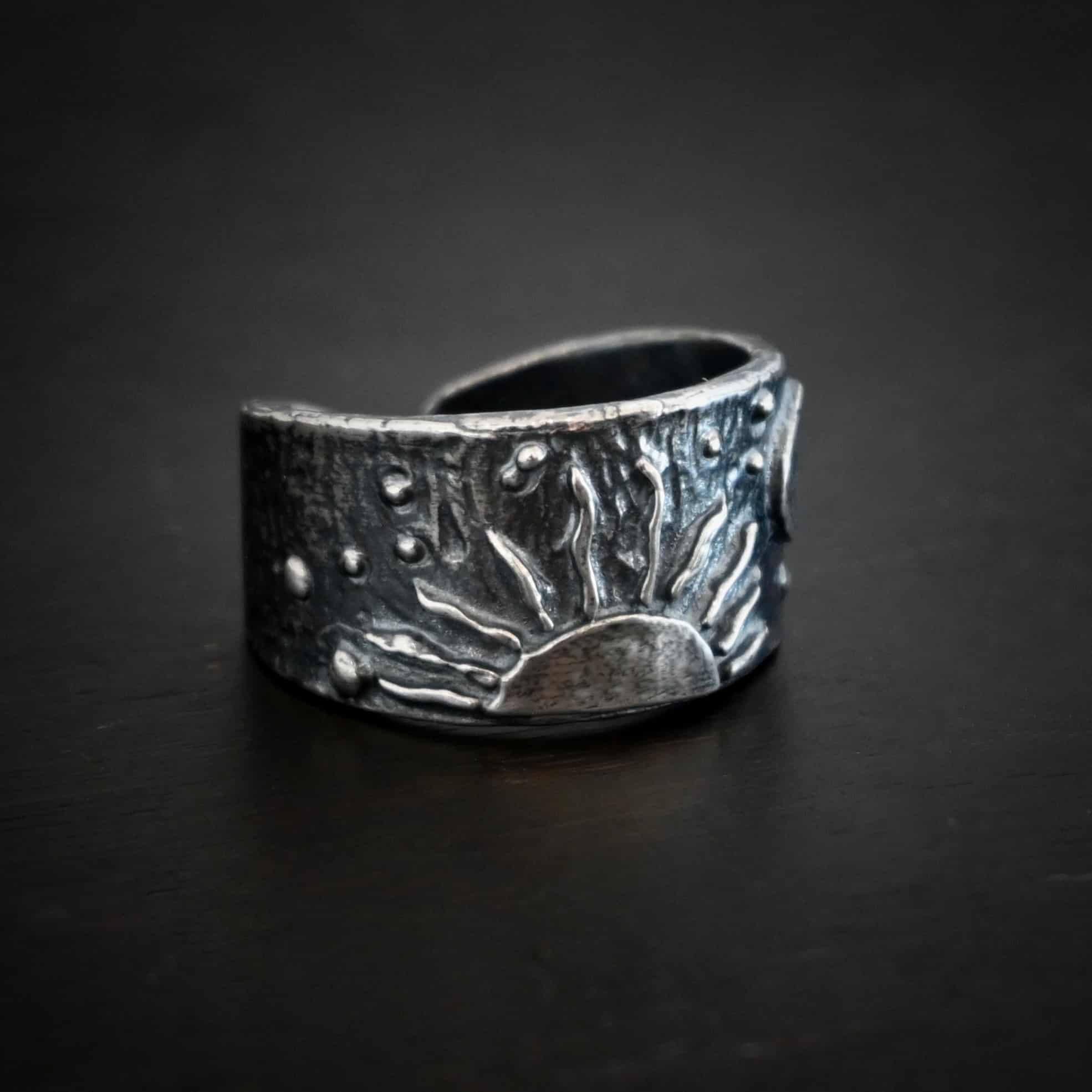 Sun Moon Stars Ring Photo of the "Sun Moon Stars" Ring. Made out of sterling silver. The bottom of the ring in the center has the top half of a rising sun. There are ten short silver lines that are wavy that go around this to signify the sun rising. There is a crescent moon on the top right side of the band. All around the band, there is silver ball detail. Ring is sitting against a dark background.