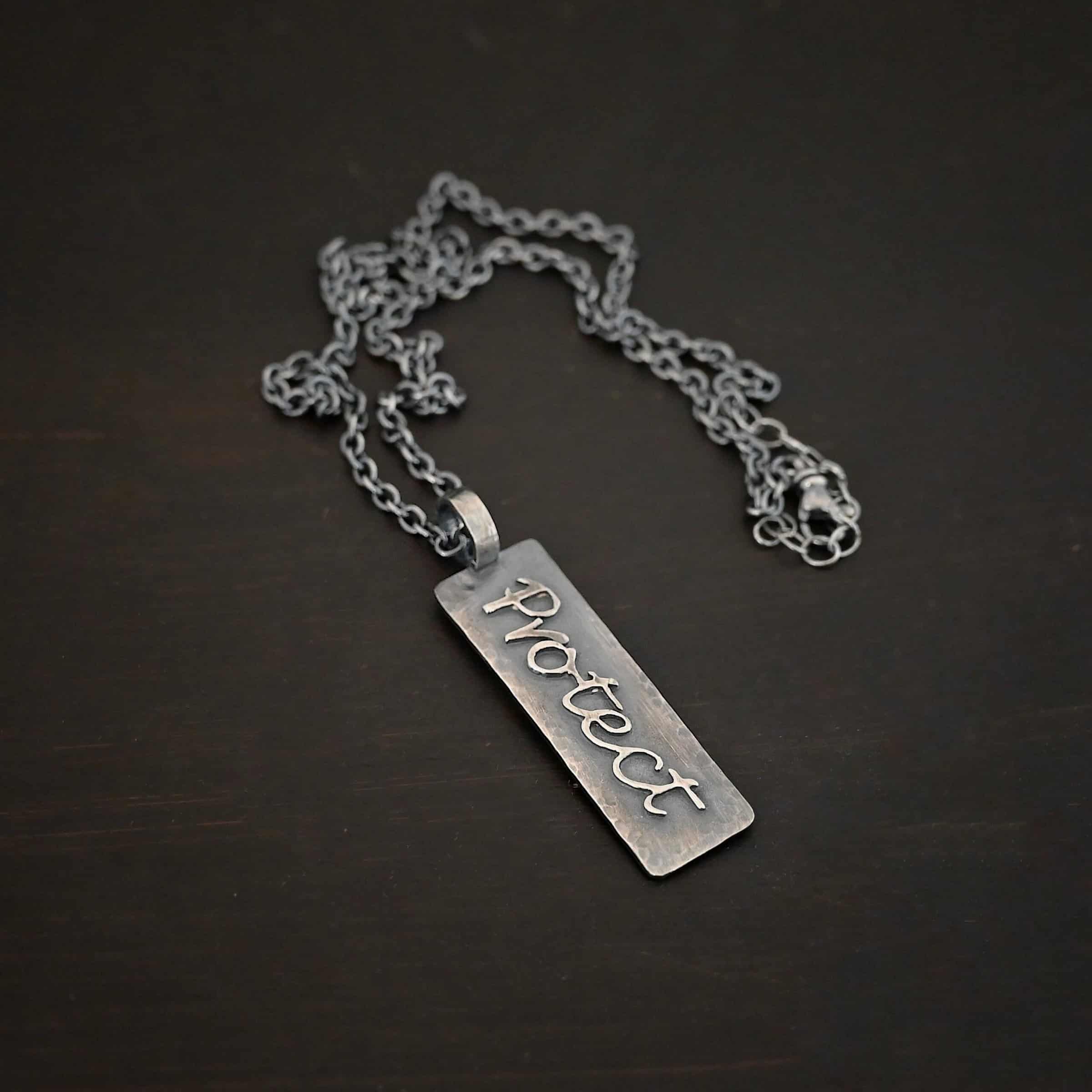 Protect Necklace Photo of the back of the "Protect" Necklace. Necklace is made out of sterling silver. The pendant is a rectangle shaped silver tag that hangs vertically. It is large. The word "Protect" is written across the entire rectangle in carved out silver and soldered to the rectangle. There is a thick jump ring soldered to the top of the pendant that the chain of the necklace goes through. Necklace is laying against a dark background.