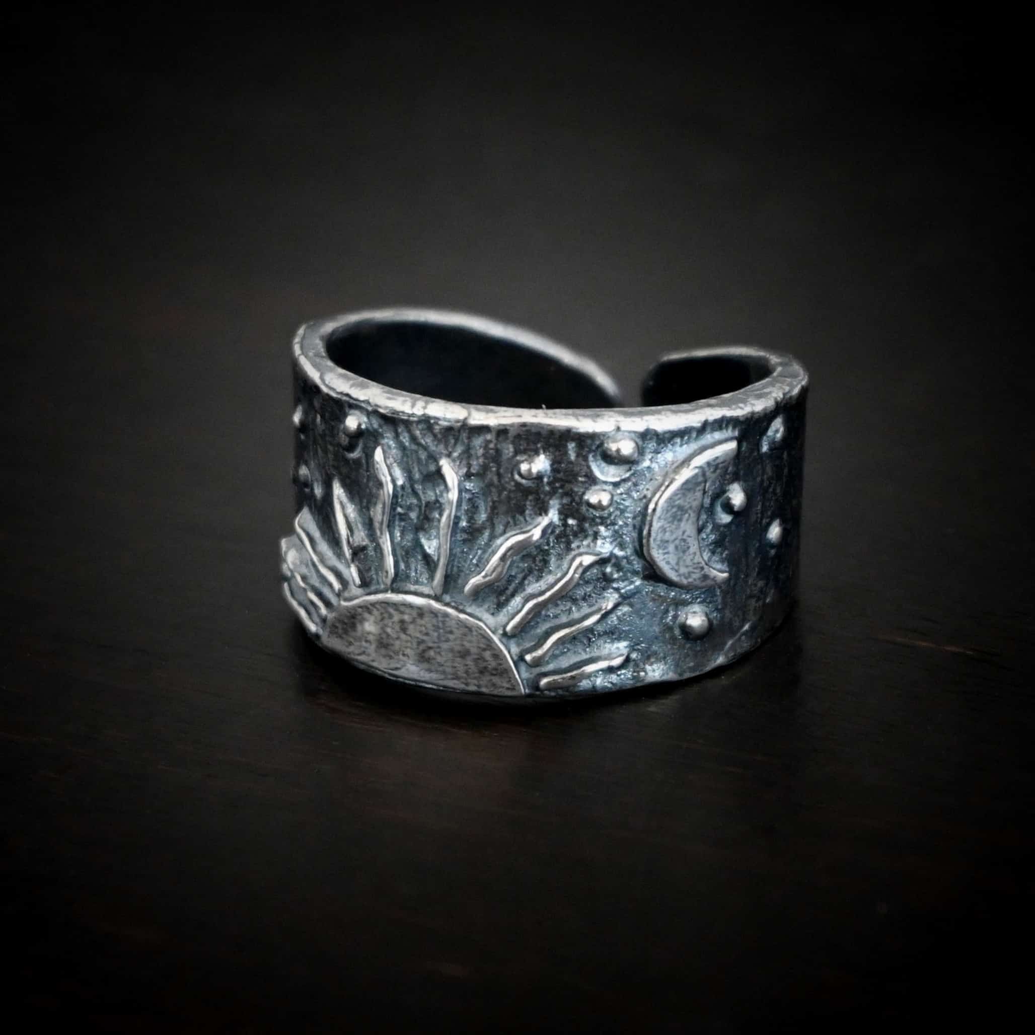 Sun Moon Stars Ring Photo of the "Sun Moon Stars" Ring. Made out of sterling silver. The bottom of the ring in the center has the top half of a rising sun. There are ten short silver lines that are wavy that go around this to signify the sun rising. There is a crescent moon on the top right side of the band. All around the band, there is silver ball detail. Ring is sitting against a dark background.