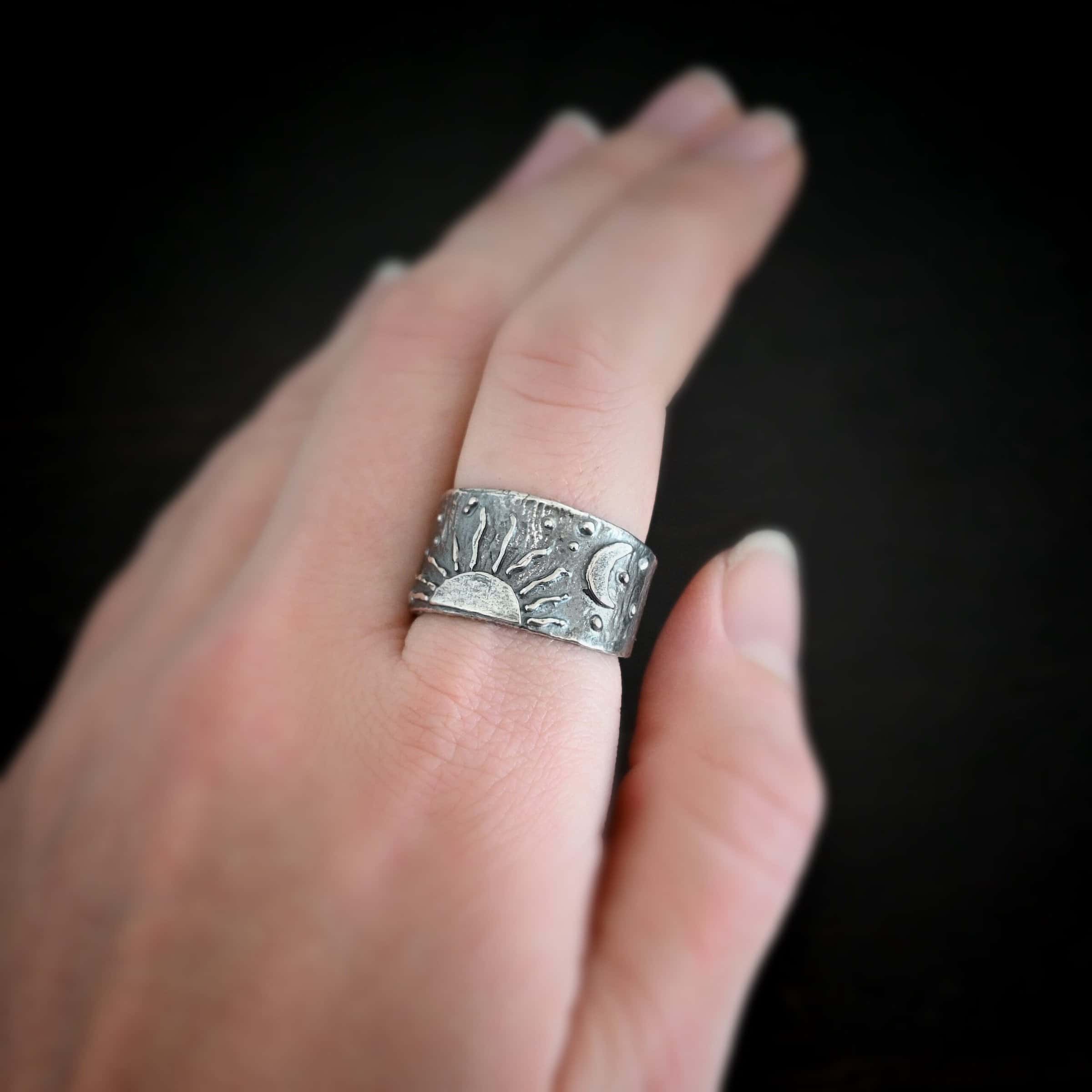 Sun Moon Stars Ring Photo of the "Sun Moon Stars" Ring. Made out of sterling silver. The bottom of the ring in the center has the top half of a rising sun. There are ten short silver lines that are wavy that go around this to signify the sun rising. There is a crescent moon on the top right side of the band. All around the band, there is silver ball detail. Ring is worn by a model and shown up close on a finger.