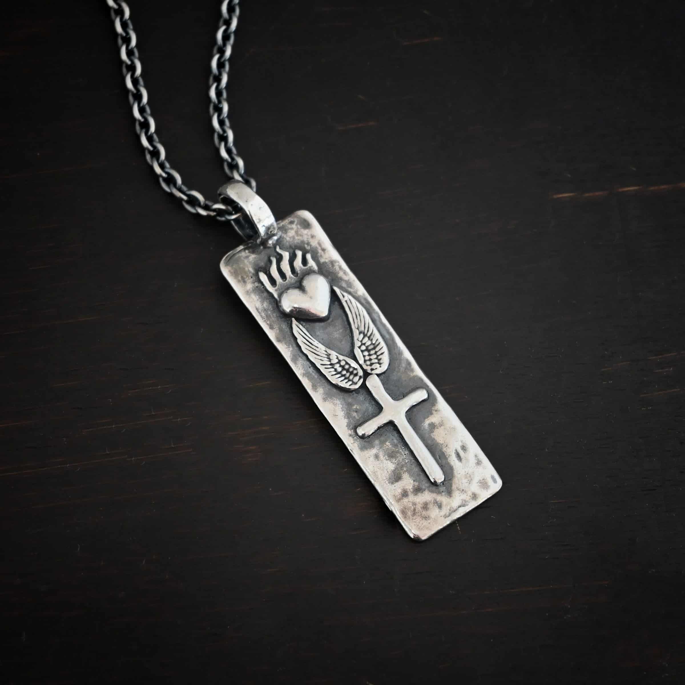 Protect Necklace Photo of the "Protect" Necklace. Necklace is made out of silver and gold. The pendant is a rectangle shaped silver tag that hangs vertically. It is large. On the top of the rectangle is a silver heart with flames dancing across the top of it. Beneath this are two silver angel wings, one on each side of the heart. Beneath the angel wings in the center of them and on the bottom of the rectangle pendant is a silver cross soldered to the pendant. There is a thick jump ring soldered to the top of the pendant that the chain of the necklace goes through. Necklace is laying against a dark background.