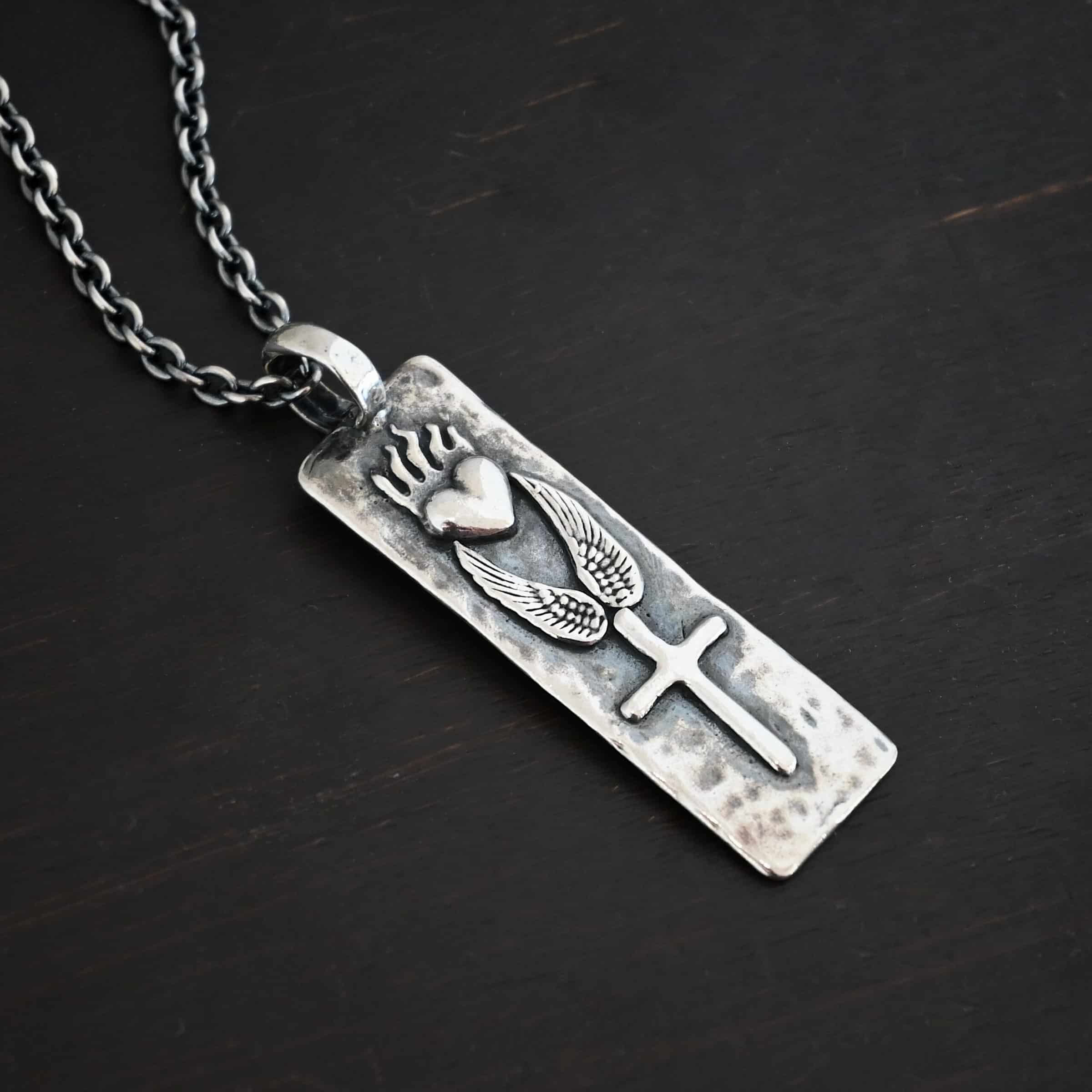 Protect Necklace Photo of the "Protect" Necklace. Necklace is made out of silver and gold. The pendant is a rectangle shaped silver tag that hangs vertically. It is large. On the top of the rectangle is a silver heart with flames dancing across the top of it. Beneath this are two silver angel wings, one on each side of the heart. Beneath the angel wings in the center of them and on the bottom of the rectangle pendant is a silver cross soldered to the pendant. There is a thick jump ring soldered to the top of the pendant that the chain of the necklace goes through. Necklace is laying against a dark background.