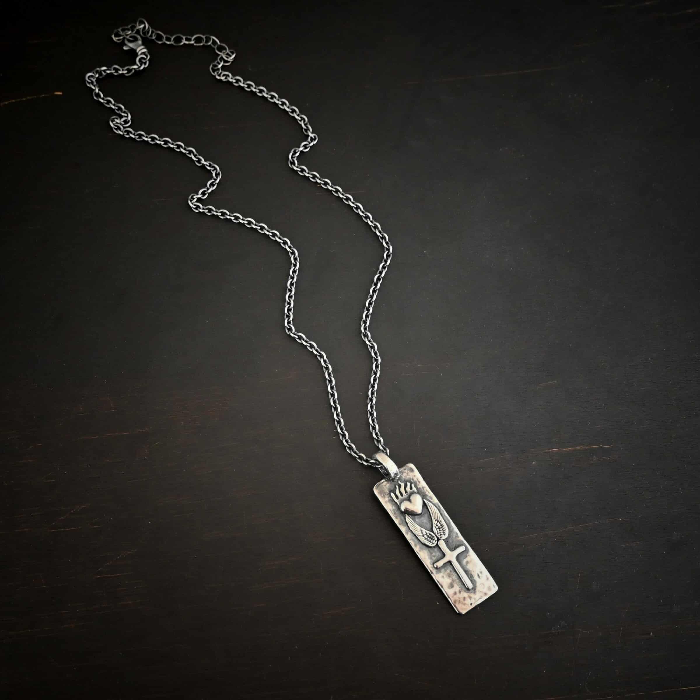 Protect Necklace Photo of the "Protect" Necklace. Necklace is made out of silver and gold. The pendant is a rectangle shaped silver tag that hangs vertically. It is large. On the top of the rectangle is a silver heart with flames dancing across the top of it. Beneath this are two silver angel wings, one on each side of the heart. Beneath the angel wings in the center of them and on the bottom of the rectangle pendant is a silver cross soldered to the pendant. There is a thick jump ring soldered to the top of the pendant that the chain of the necklace goes through. Necklace is laying against a dark background.