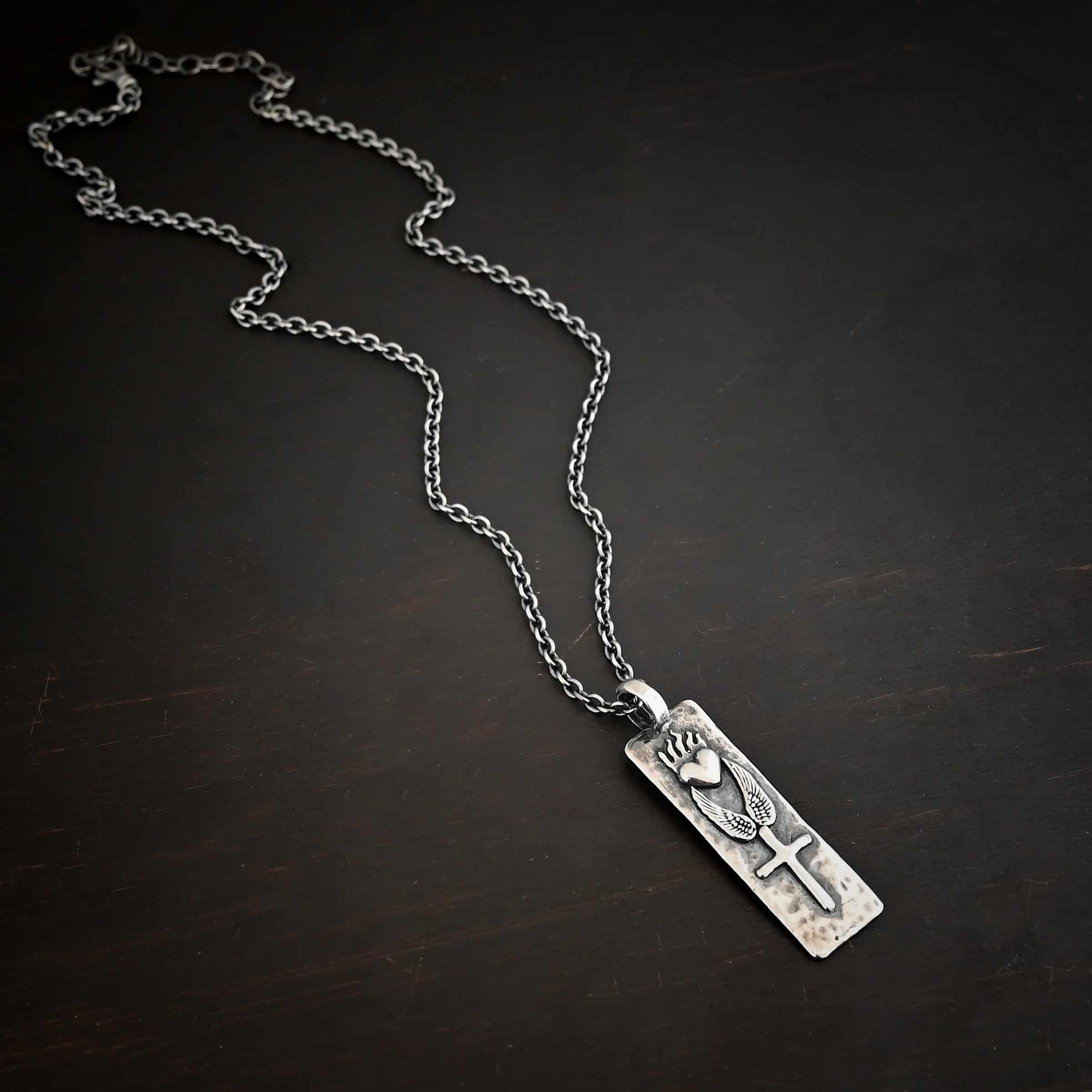 Protect Necklace Photo of the "Protect" Necklace. Necklace is made out of silver and gold. The pendant is a rectangle shaped silver tag that hangs vertically. It is large. On the top of the rectangle is a silver heart with flames dancing across the top of it. Beneath this are two silver angel wings, one on each side of the heart. Beneath the angel wings in the center of them and on the bottom of the rectangle pendant is a silver cross soldered to the pendant. There is a thick jump ring soldered to the top of the pendant that the chain of the necklace goes through. Necklace is laying against a dark background.