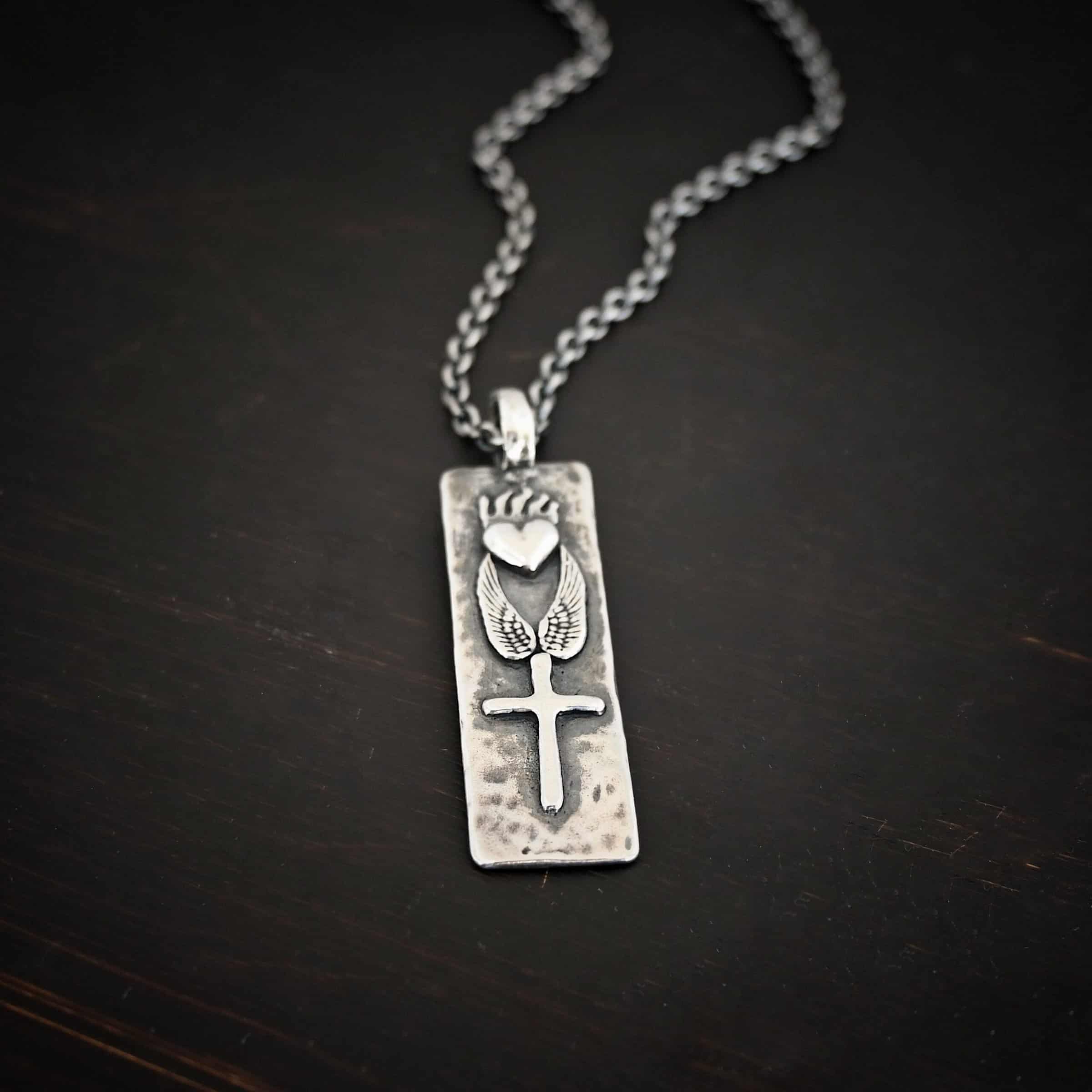 Protect Necklace Photo of the "Protect" Necklace. Necklace is made out of silver and gold. The pendant is a rectangle shaped silver tag that hangs vertically. It is large. On the top of the rectangle is a silver heart with flames dancing across the top of it. Beneath this are two silver angel wings, one on each side of the heart. Beneath the angel wings in the center of them and on the bottom of the rectangle pendant is a silver cross soldered to the pendant. There is a thick jump ring soldered to the top of the pendant that the chain of the necklace goes through. Necklace is laying against a dark background.