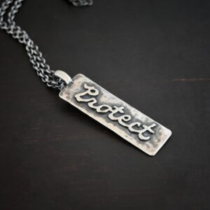 Photo of the back of the "Protect" Necklace. Necklace is made out of sterling silver. The pendant is a rectangle shaped silver tag that hangs vertically. It is large. The word "Protect" is written across the entire rectangle in carved out silver and soldered to the rectangle. There is a thick jump ring soldered to the top of the pendant that the chain of the necklace goes through. Necklace is laying against a dark background.