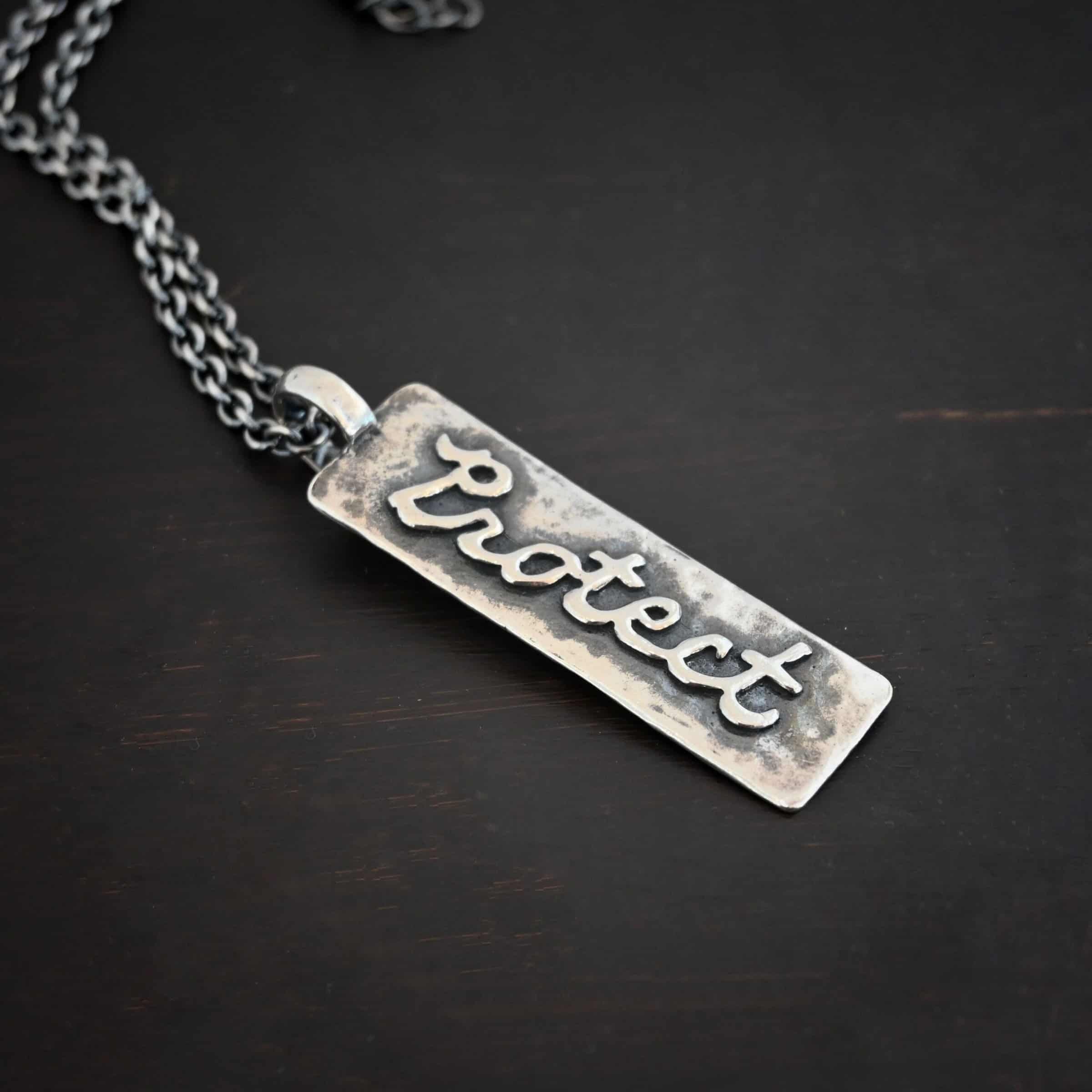 Protect Necklace Photo of the back of the "Protect" Necklace. Necklace is made out of sterling silver. The pendant is a rectangle shaped silver tag that hangs vertically. It is large. The word "Protect" is written across the entire rectangle in carved out silver and soldered to the rectangle. There is a thick jump ring soldered to the top of the pendant that the chain of the necklace goes through. Necklace is laying against a dark background.