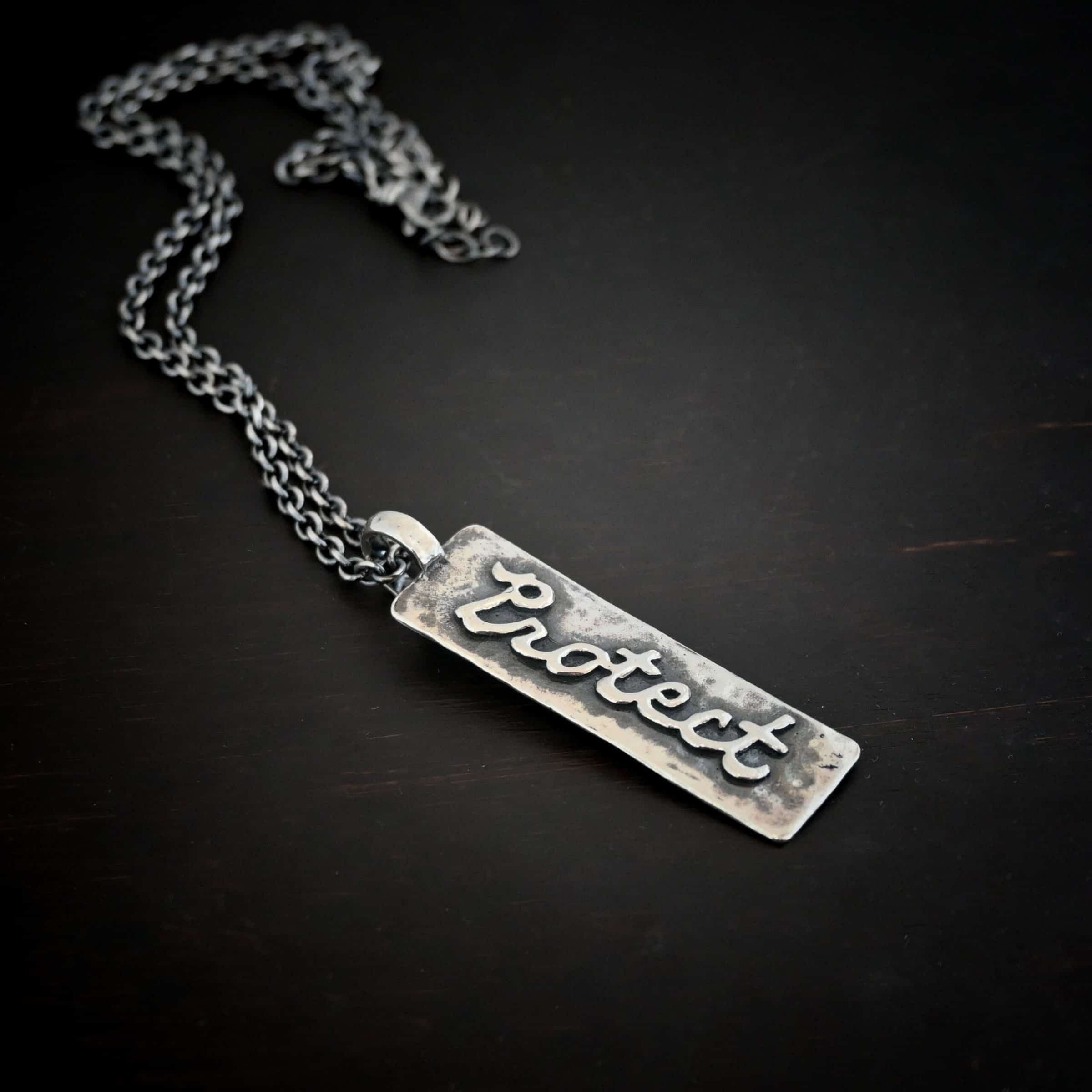 Protect Necklace Photo of the back of the "Protect" Necklace. Necklace is made out of sterling silver. The pendant is a rectangle shaped silver tag that hangs vertically. It is large. The word "Protect" is written across the entire rectangle in carved out silver and soldered to the rectangle. There is a thick jump ring soldered to the top of the pendant that the chain of the necklace goes through. Necklace is laying against a dark background.