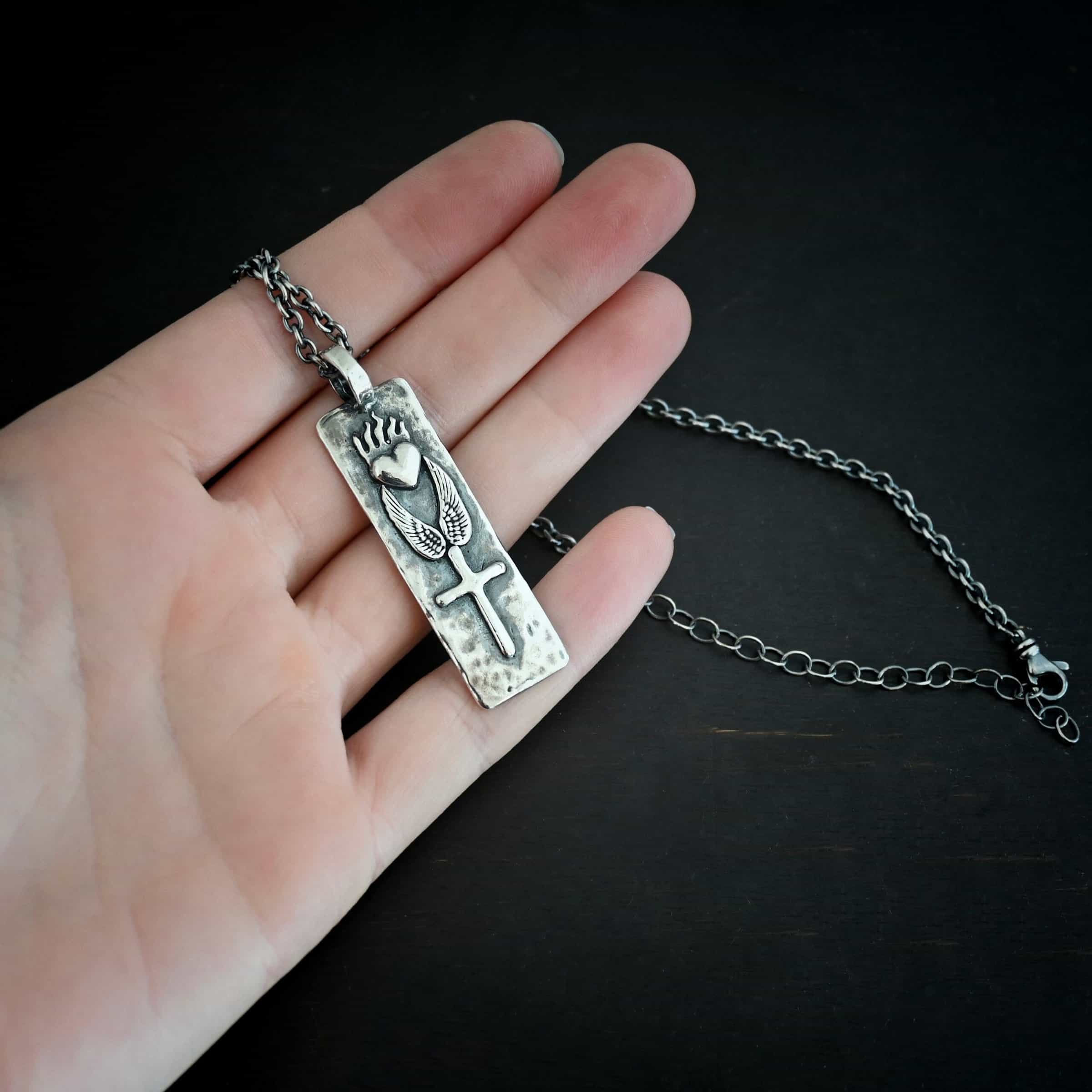 Protect Necklace Photo of the "Protect" Necklace. Necklace is made out of silver and gold. The pendant is a rectangle shaped silver tag that hangs vertically. It is large. On the top of the rectangle is a silver heart with flames dancing across the top of it. Beneath this are two silver angel wings, one on each side of the heart. Beneath the angel wings in the center of them and on the bottom of the rectangle pendant is a silver cross soldered to the pendant. There is a thick jump ring soldered to the top of the pendant that the chain of the necklace goes through. Necklace is laying against the fingertips of a hand.