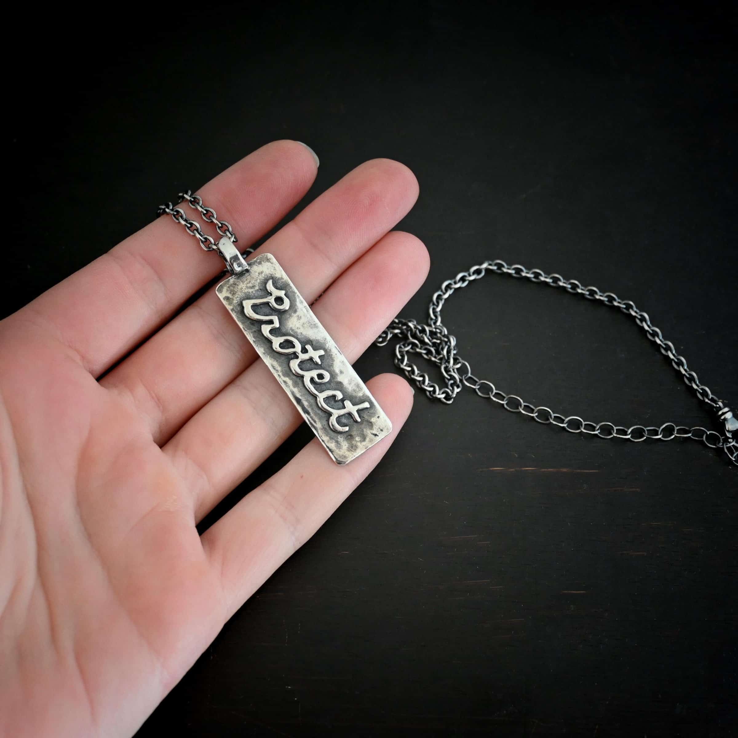 Protect Necklace Photo of the back of the "Protect" Necklace. Necklace is made out of sterling silver. The pendant is a rectangle shaped silver tag that hangs vertically. It is large. The word "Protect" is written across the entire rectangle in carved out silver and soldered to the rectangle. There is a thick jump ring soldered to the top of the pendant that the chain of the necklace goes through. Necklace is laying against the fingertips of a hand to demonstrate size.