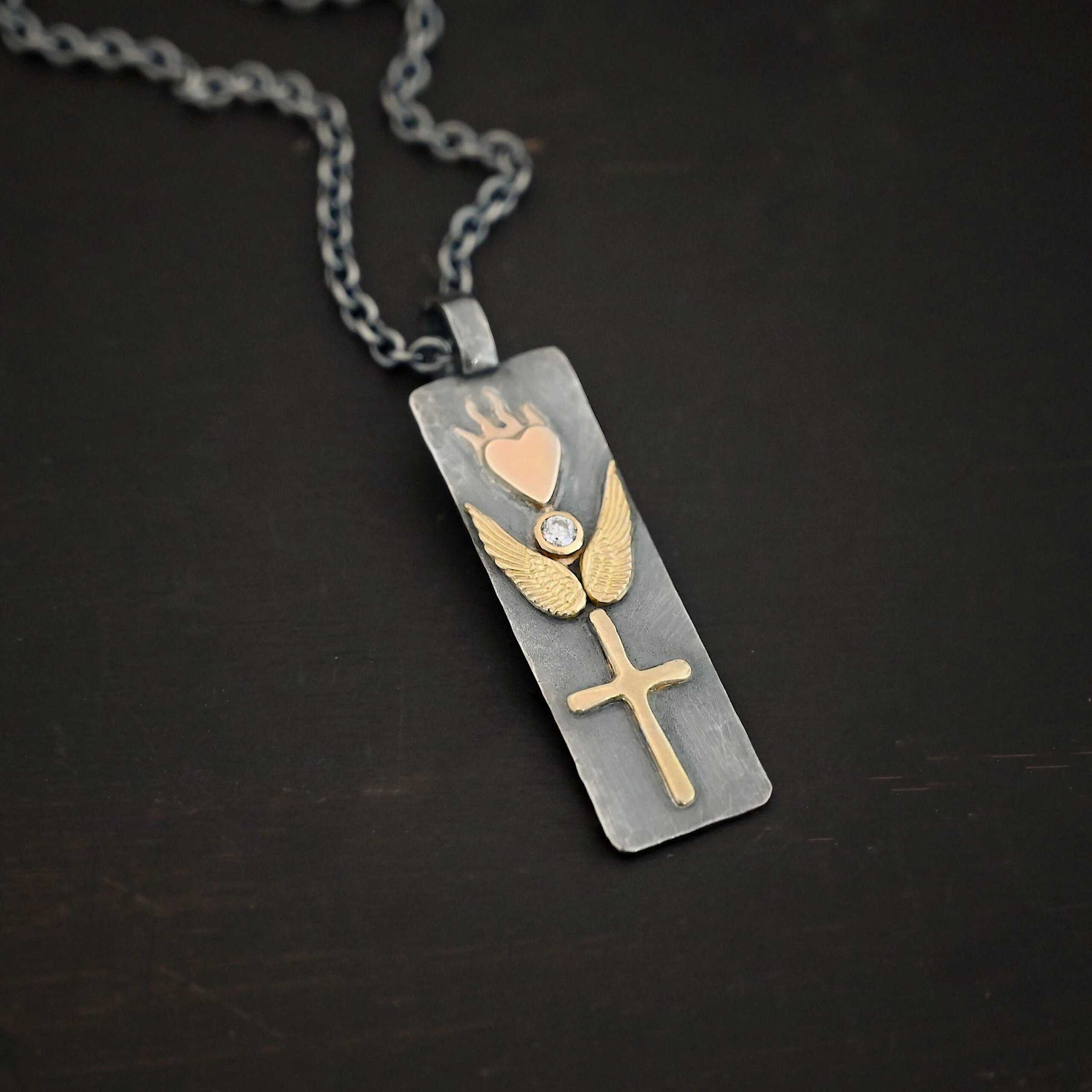 Protect Necklace Photo of the "Protect" Necklace. Necklace is made out of silver and gold. The pendant is a rectangle shaped silver tag that hangs vertically. It is large. On the top of the rectangle is a gold heart with gold flames dancing across the top of it. On the bottom, beneath the heart is a diamond that is set in gold. On each side of the diamond is a gold angel wing. Beneath the gold angel wings in the center of them and on the bottom of the rectangle pendant is a gold cross. There is a thick jump ring soldered to the top of the pendant that the chain of the necklace goes through. Necklace is laying against a dark background.