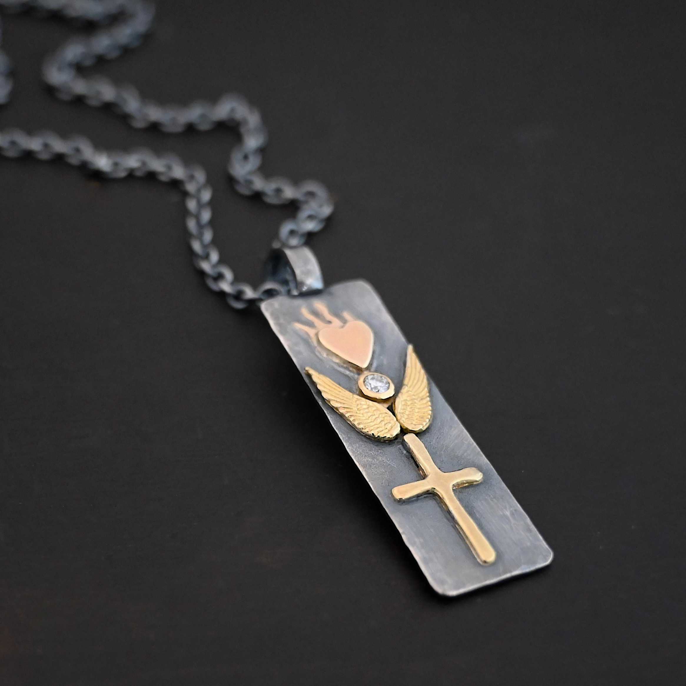 Protect Necklace Photo of the "Protect" Necklace. Necklace is made out of silver and gold. The pendant is a rectangle shaped silver tag that hangs vertically. It is large. On the top of the rectangle is a gold heart with gold flames dancing across the top of it. On the bottom, beneath the heart is a diamond that is set in gold. On each side of the diamond is a gold angel wing. Beneath the gold angel wings in the center of them and on the bottom of the rectangle pendant is a gold cross. There is a thick jump ring soldered to the top of the pendant that the chain of the necklace goes through. Necklace is laying against a dark background.
