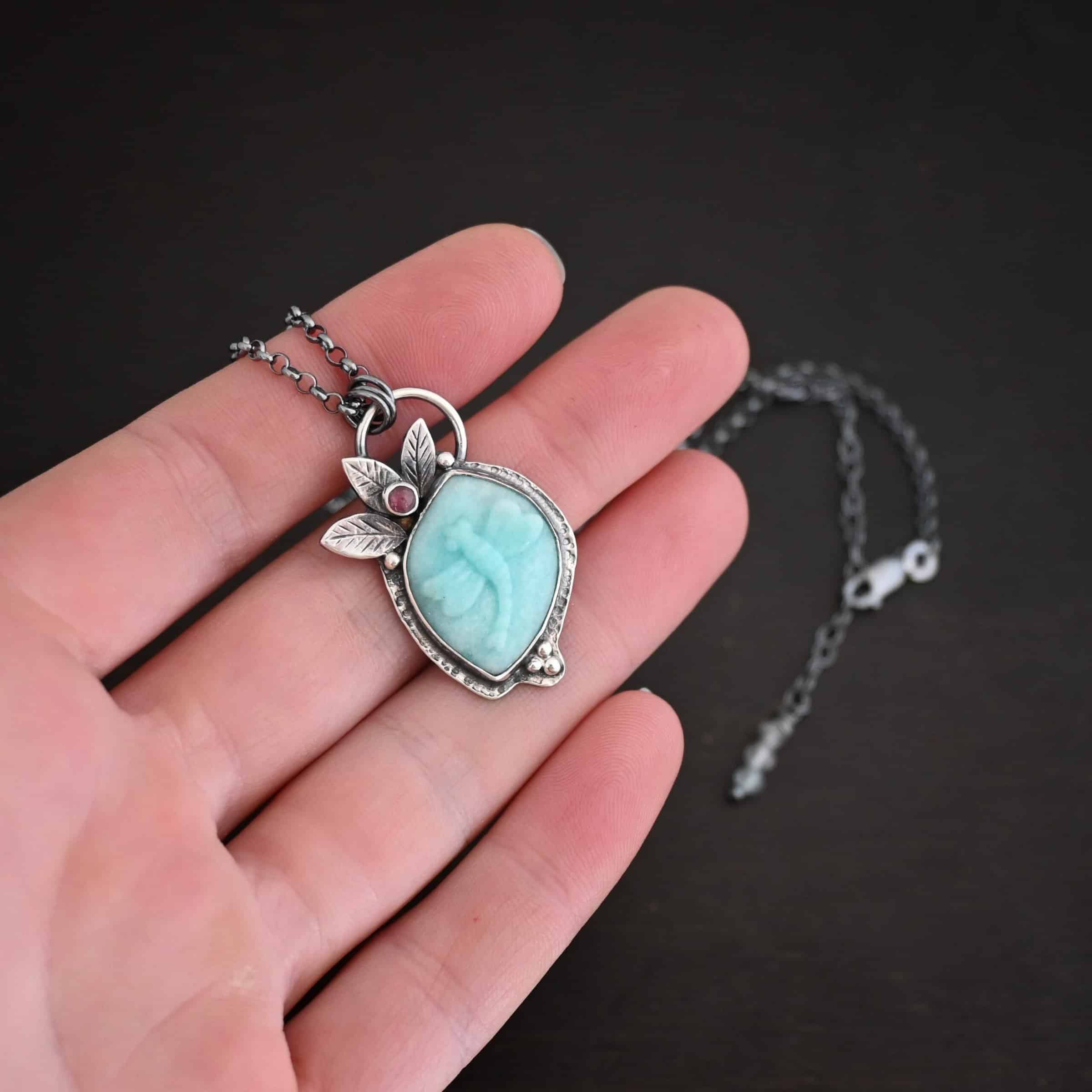 Close up of the "Libelle" Necklace. Made out of amazonite and pink tourmaline. The amazonite is an oval shape, but with pointed ends on it. It is a light blue color and has a dragonfly carved into it. There are three silver leaves in the top left corner of the necklace, and the pink tourmaline is in the middle of the leaves. There are five silver dots on the necklace. One on each side of the leaves, and three in the bottom right corner of the necklace. Pendant has a loop on the back of it to attach it to jump rings that are hanging on a chain that is sterling silver and oxidized. Pendant is laying against the fingertips of a hand.