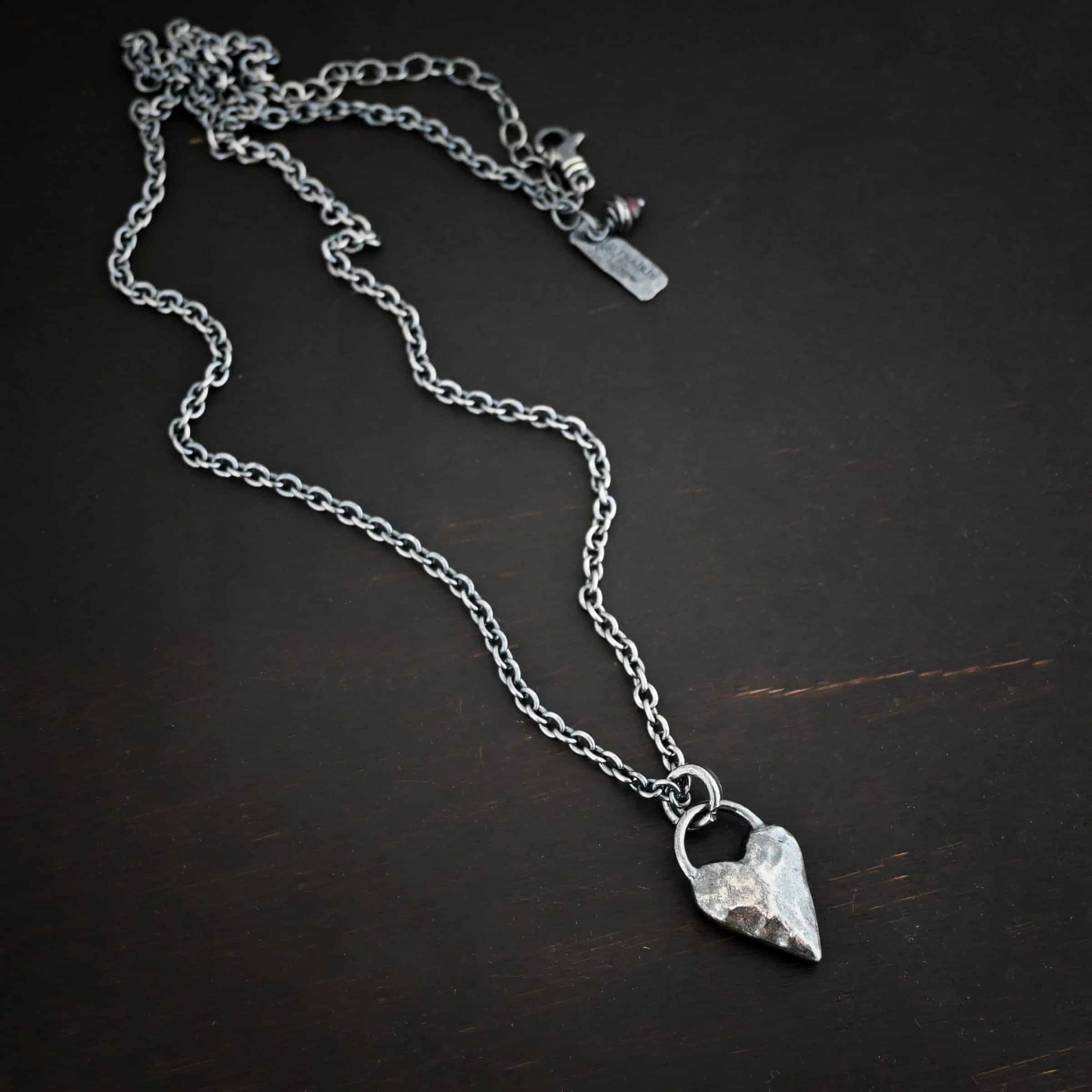 Photo of the "Heart Throb" Necklace. Made out of sterling silver. There is a thick, solid sterling silver heart that is a medium size for the pendant. It is hammered and oxidized. There is a thick semicircle that is soldered on both sides of the heart on the top. Attached to the semicircle is a thick jump ring that the chain of the necklace goes through. Necklace is lying against a dark background.