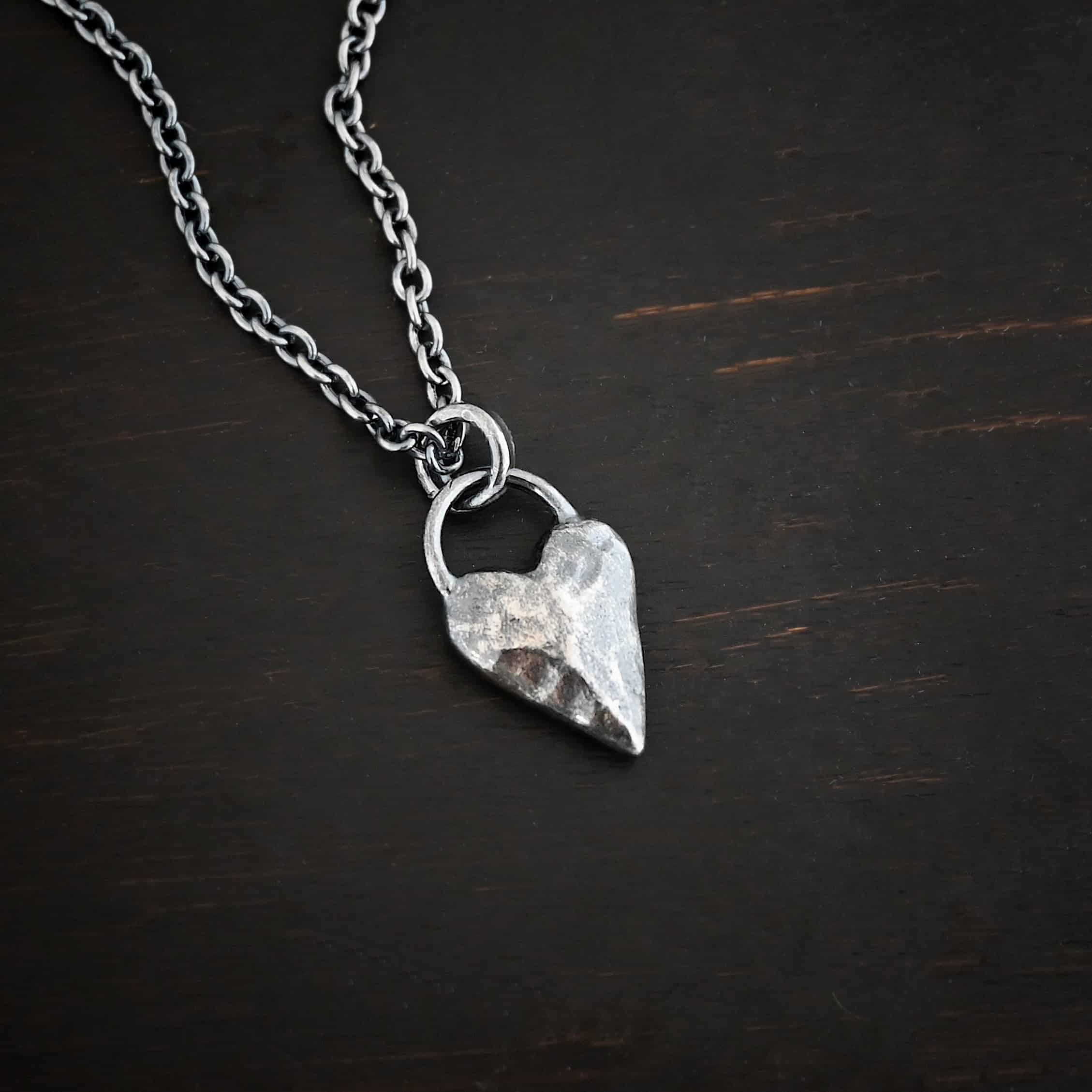 Photo of the "Heart Throb" Necklace. Made out of sterling silver. There is a thick, solid sterling silver heart that is a medium size for the pendant. It is hammered and oxidized. There is a thick semicircle that is soldered on both sides of the heart on the top. Attached to the semicircle is a thick jump ring that the chain of the necklace goes through. Necklace is lying against a dark background.