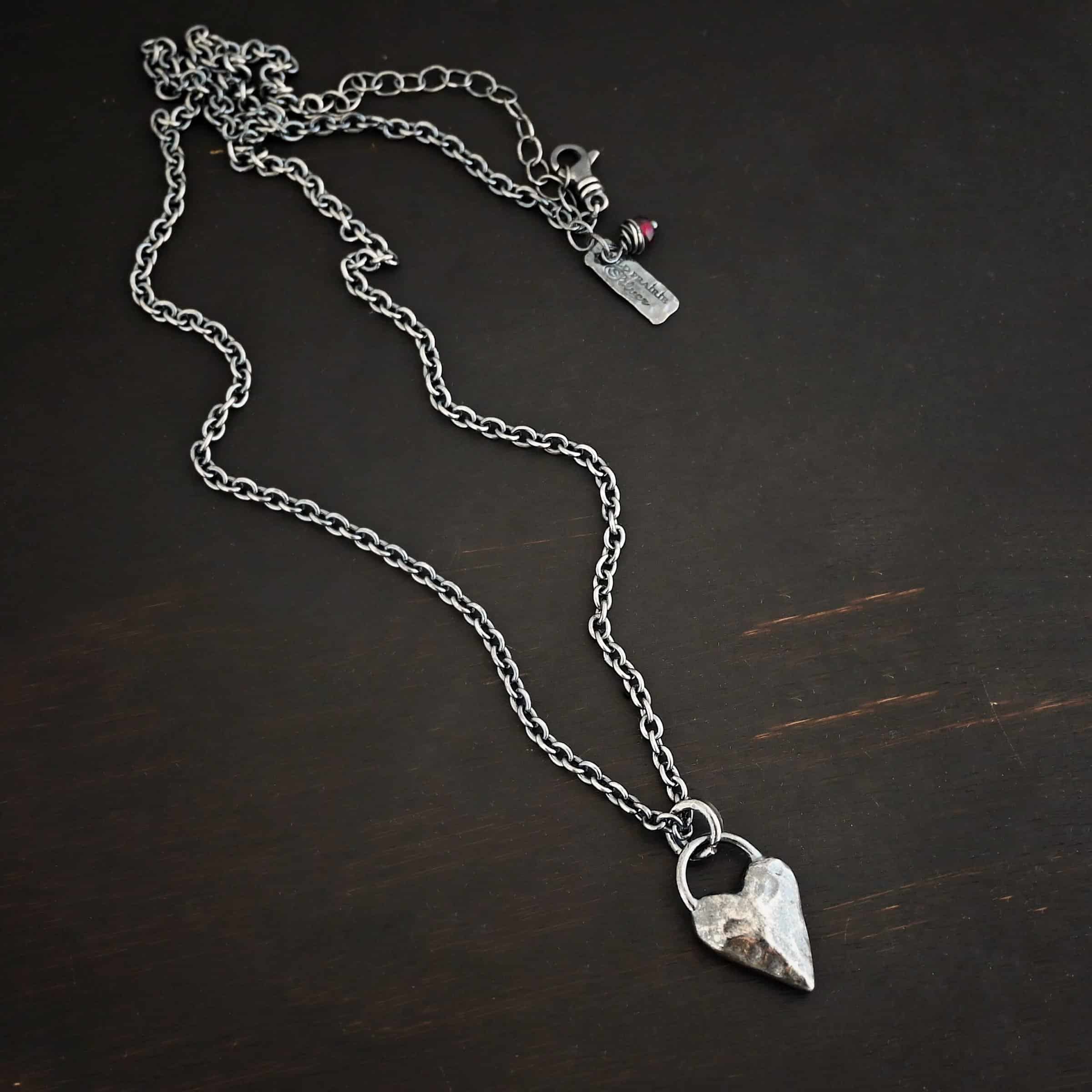 Photo of the "Heart Throb" Necklace. Made out of sterling silver. There is a thick, solid sterling silver heart that is a medium size for the pendant. It is hammered and oxidized. There is a thick semicircle that is soldered on both sides of the heart on the top. Attached to the semicircle is a thick jump ring that the chain of the necklace goes through. Necklace is lying against a dark background.