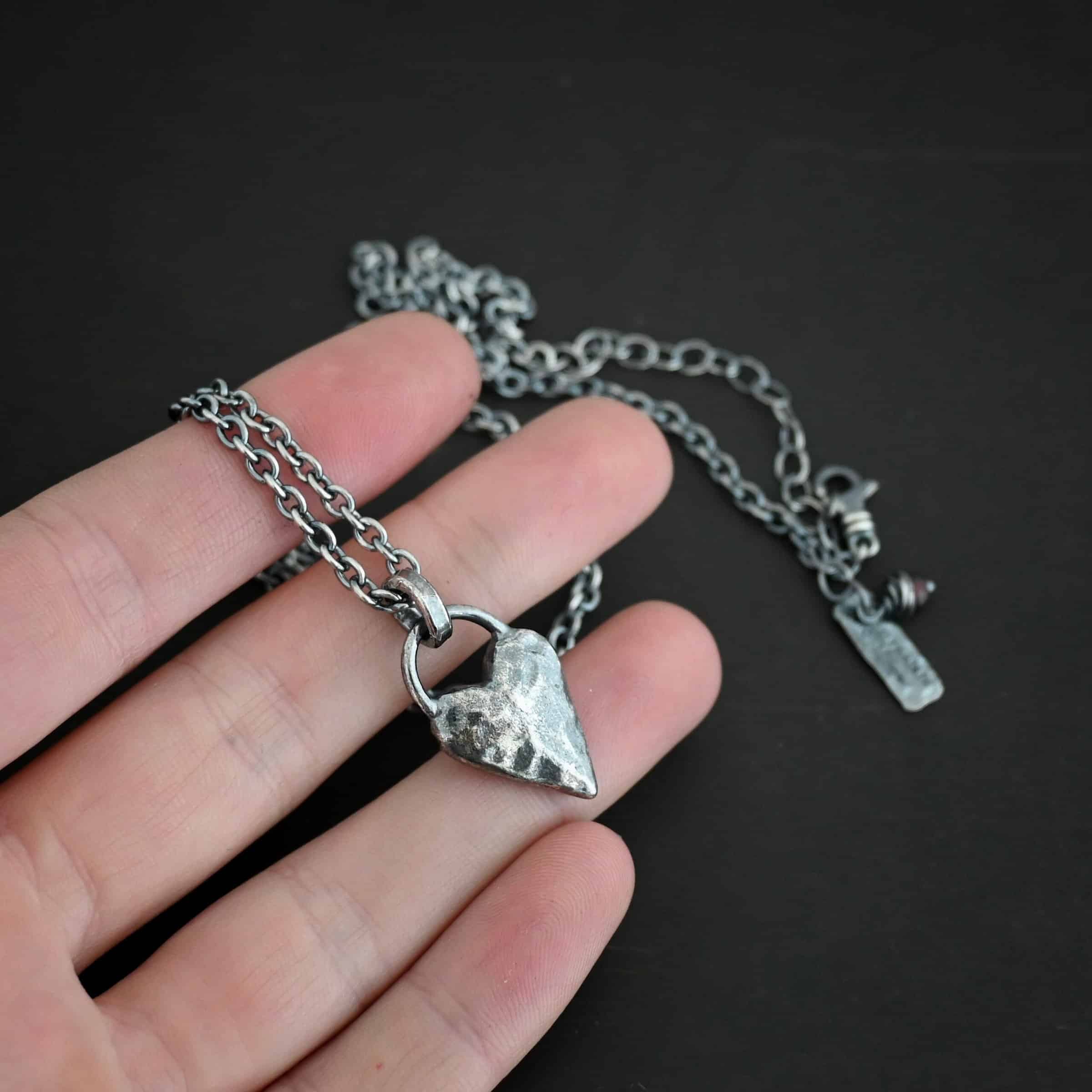 Photo of the "Heart Throb" Necklace. Made out of sterling silver. There is a thick, solid sterling silver heart that is a medium size for the pendant. It is hammered and oxidized. There is a thick semicircle that is soldered on both sides of the heart on the top. Attached to the semicircle is a thick jump ring that the chain of the necklace goes through. Necklace is lying against the fingertips of a hand.