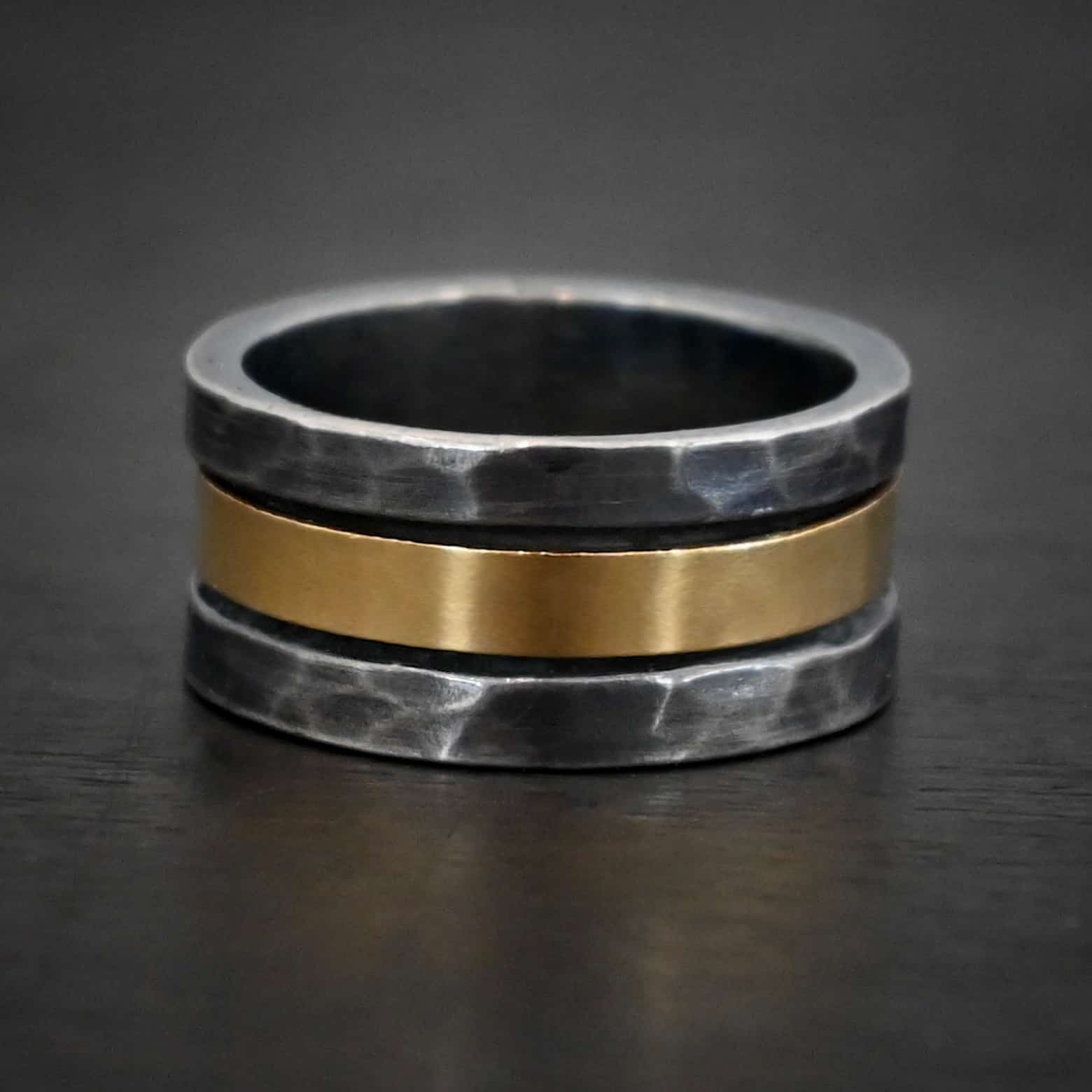 Photo of the "Tough As Nails" Wedding Band. Made out of sterling silver and gold. The top and bottom of the band are hammered and oxidized sterling silver. There is a thick band of gold going through the center and around the entire ring. There is a small gap before the gold starts and ends in the ring. The gold is smooth. Ring is sitting against a dark background.