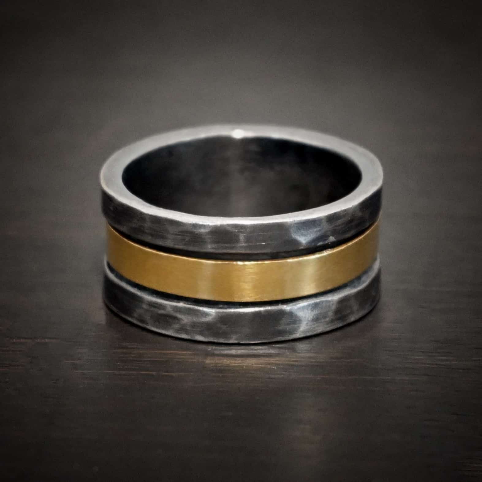 Photo of the "Tough As Nails" Wedding Band. Made out of sterling silver and gold. The top and bottom of the band are hammered and oxidized sterling silver. There is a thick band of gold going through the center and around the entire ring. There is a small gap before the gold starts and ends in the ring. The gold is smooth. Ring is sitting against a dark background.