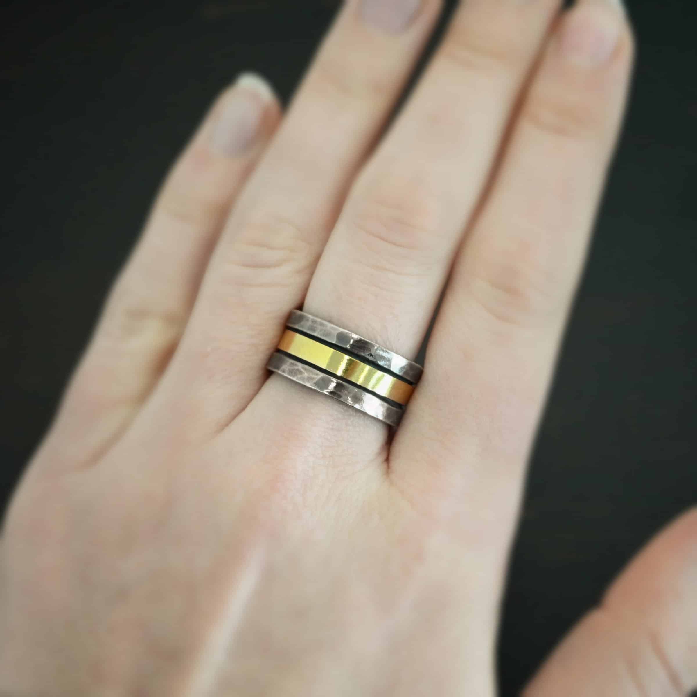 Photo of the "Tough As Nails" Wedding Band. Made out of sterling silver and gold. The top and bottom of the band are hammered and oxidized sterling silver. There is a thick band of gold going through the center and around the entire ring. There is a small gap before the gold starts and ends in the ring. The gold is smooth. Ring is worn by a model.