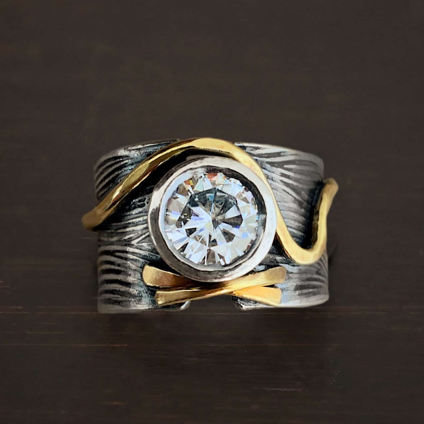 Endless Love Photo of the "Endless Love" Ring. Made out of sterling silver, gold, and moissanite. The center of the ring does not connect. There is a large moissanite that is set in silver in the center of the ring that connects both sides of the band. There are two small lines of gold beneath the moissanite that cross to create an "x" shape. Above the moissanite is another line of gold that curves to go around the moissanite, and then dips down and up again as it goes across the ring. The rest of the silver band is engraved with wavy lines. Ring is set against a dark background.