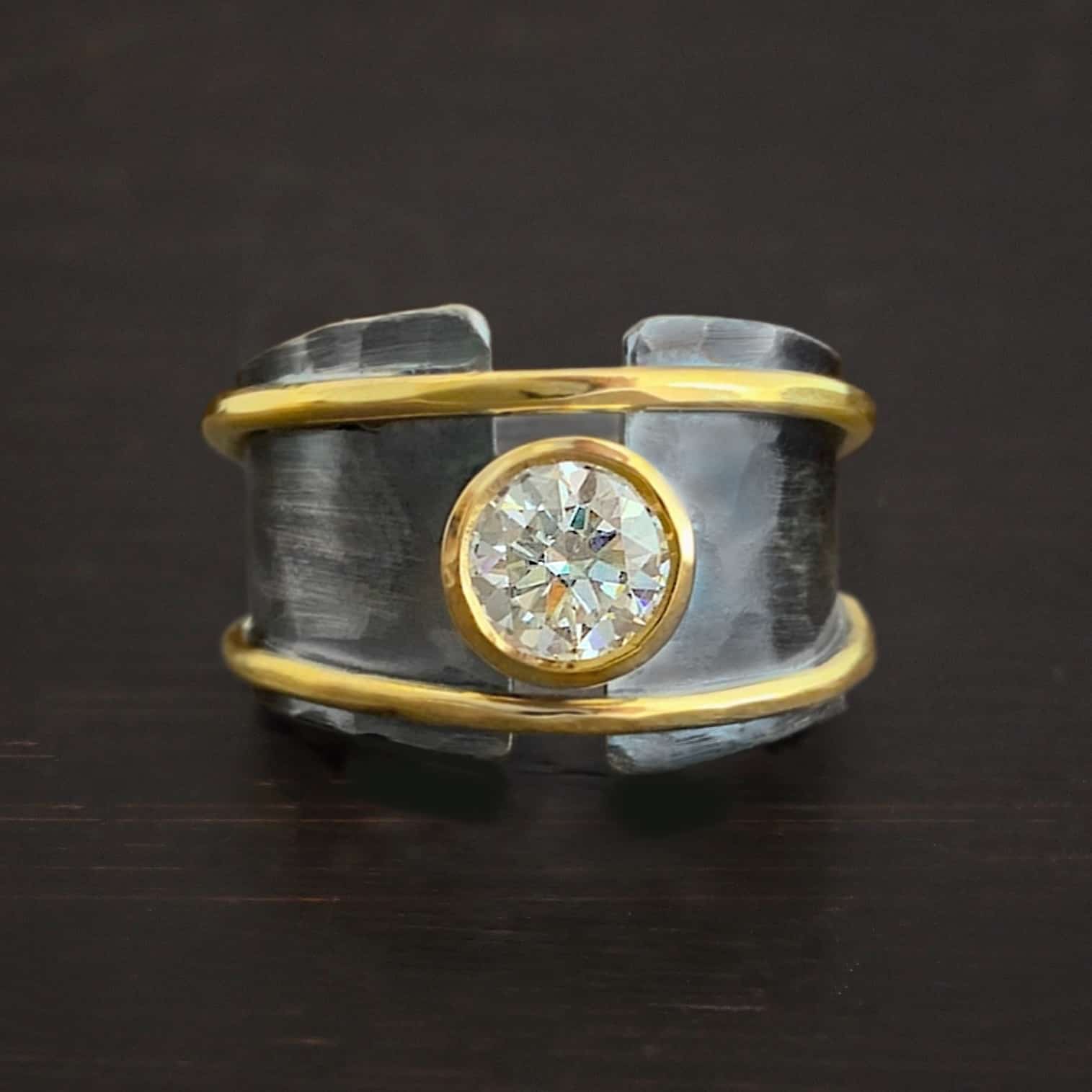 Banded Together Ring Photo of the "Banded Together" Ring. Made out of sterling silver, gold, and moissanite. The ring band does not fully connect. It is thick and hammered. There is a moissanite in the center that connects both sides. It is set in gold. There is a thin band of gold on top and on the bottom of the ring. They both go fully around and connect both sides of the ring as well. Ring is set against a dark background.