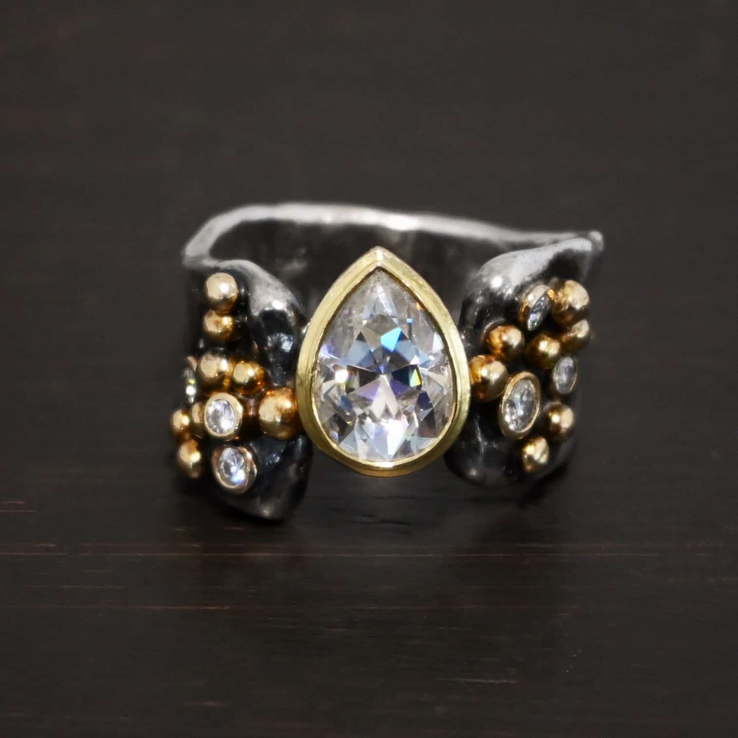 Head Over Boots Ring Photo of the "Head Over Boots" Ring. Made out of sterling silver, gold, and moissanites. There is a large gap in the center of the ring band. The ring band is also uneven, so one side of the ring is shorter than the other side. There is a lot of gold ball detail on both sides of the ring. Each side of the ring has three moissanites set in gold. The center moissanite is a pear shape and set in gold. It is set in the gap of the ring band, and connects the band together. The center moissanite is connected on both sides to each side of the ring band. Ring is set against a dark background.