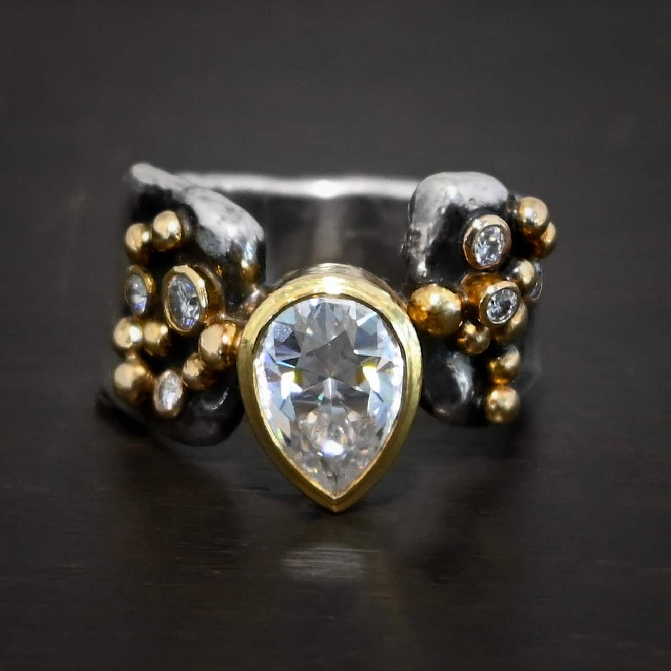 Head Over Boots Ring Photo of the "Head Over Boots" Ring. Made out of sterling silver, gold, and moissanites. There is a large gap in the center of the ring band. The ring band is also uneven, so one side of the ring is shorter than the other side. There is a lot of gold ball detail on both sides of the ring. Each side of the ring has three moissanites set in gold. The center moissanite is a pear shape and set in gold. It is set in the gap of the ring band, and connects the band together. The center moissanite is connected on both sides to each side of the ring band. Ring is set against a dark background.