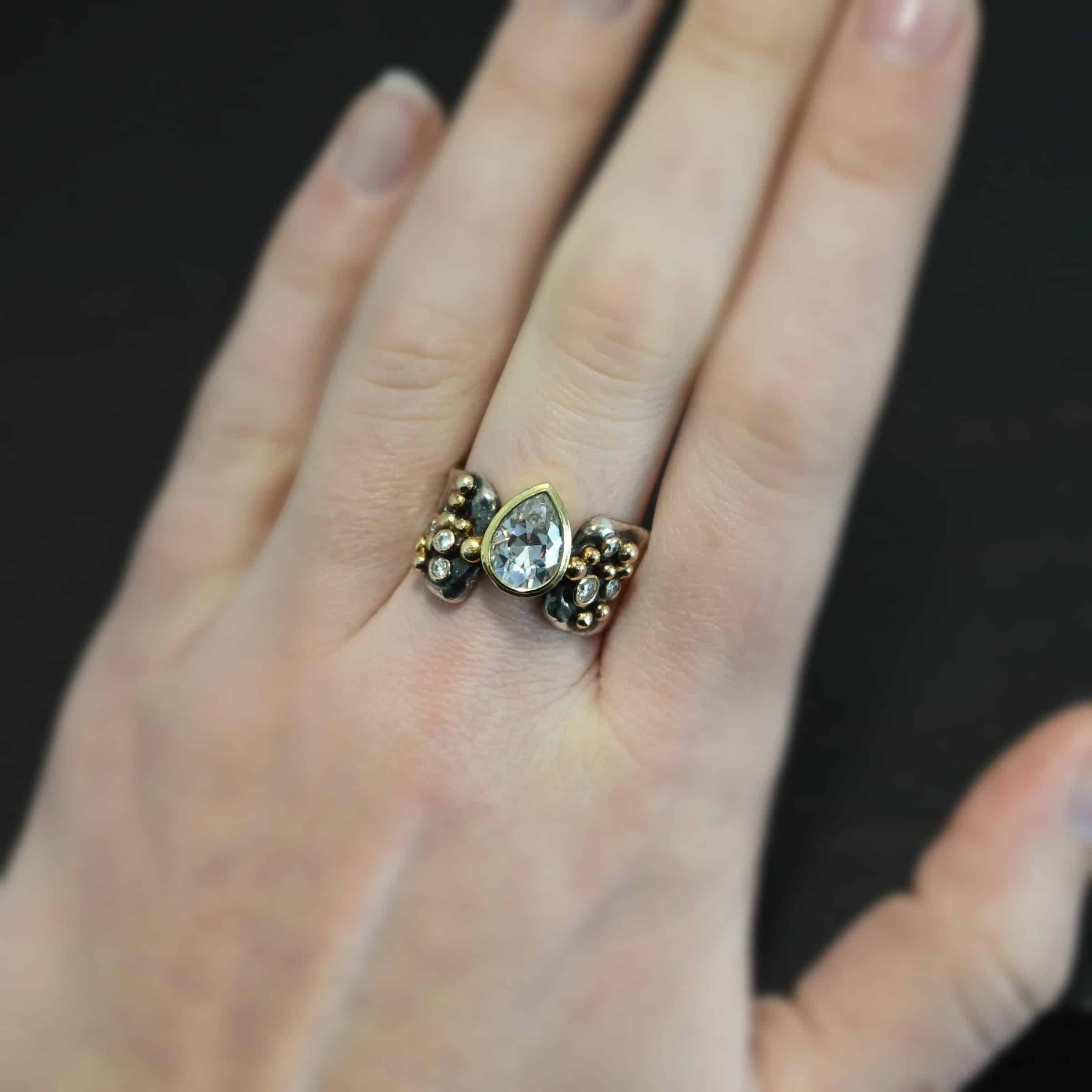 Head Over Boots Ring Photo of the "Head Over Boots" Ring. Made out of sterling silver, gold, and moissanites. There is a large gap in the center of the ring band. The ring band is also uneven, so one side of the ring is shorter than the other side. There is a lot of gold ball detail on both sides of the ring. Each side of the ring has three moissanites set in gold. The center moissanite is a pear shape and set in gold. It is set in the gap of the ring band, and connects the band together. The center moissanite is connected on both sides to each side of the ring band. Ring is worn by a model.