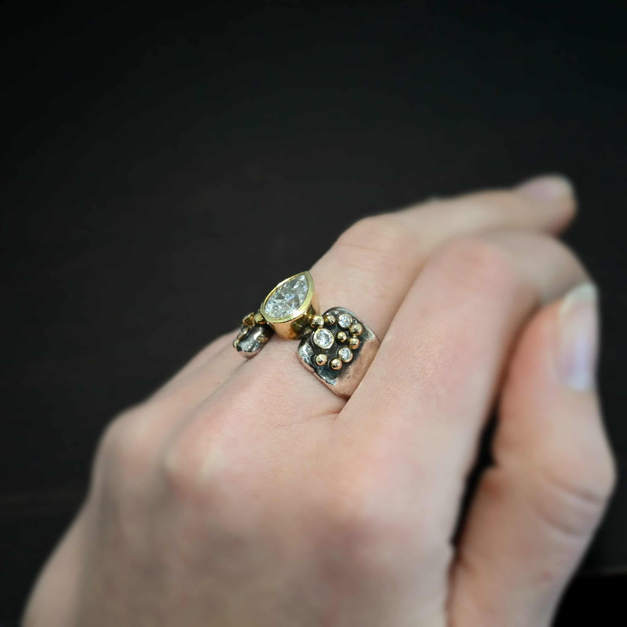 Head Over Boots Ring Photo of the "Head Over Boots" Ring. Made out of sterling silver, gold, and moissanites. There is a large gap in the center of the ring band. The ring band is also uneven, so one side of the ring is shorter than the other side. There is a lot of gold ball detail on both sides of the ring. Each side of the ring has three moissanites set in gold. The center moissanite is a pear shape and set in gold. It is set in the gap of the ring band, and connects the band together. The center moissanite is connected on both sides to each side of the ring band. Ring is worn by a model.