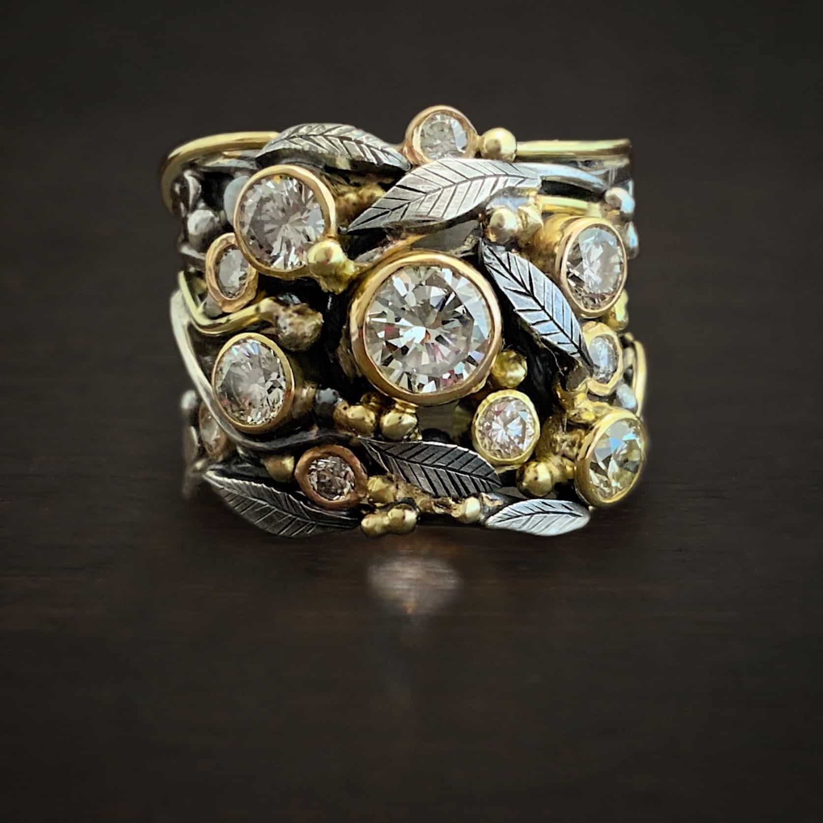Photo of the "Enchanted" Ring. Made out of sterling silver, gold, and moissanites. There are nine moissanites scattered around the center and sides of the ring. The largest one is in the center. There are silver leaves and vines around the ring. Each stone is set in gold, and there are multiple gold balls filling in all of the gaps of the ring.