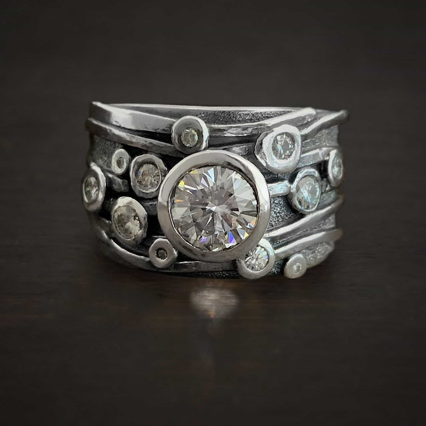 Photo of the "Cross My Heart" Ring. Made out of sterling silver, moissanite, and diamonds. There is a large moissanite in the center of the ring. There are diamonds scattered around the rest of the ring. There are five, thicker lines of silver on the ring. One line starts at the top, and the others follow to the bottom of the ring.