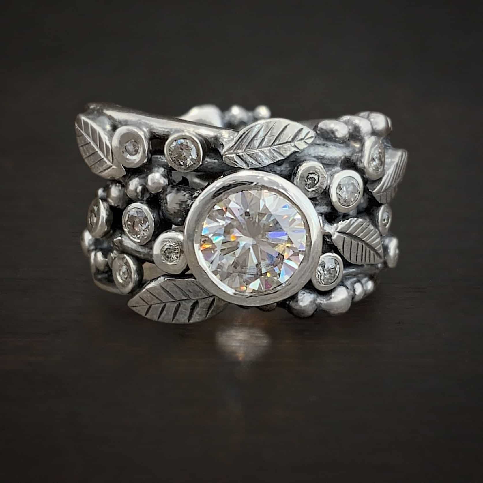 Photo of the "Enchanted" Ring. There is a large moissanite in the center of the band. There are diamonds scattered all around the rest of the ring. There are silver leaves around the ring as well. The rest of the ring has multiple balls of silver scattered all around the ring. Ring is set against a dark background.