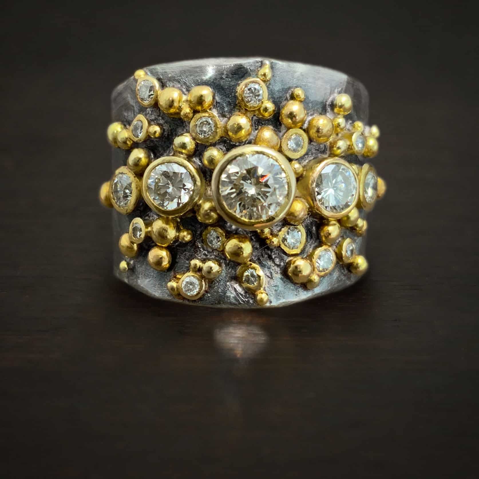 Photo of the "Starstruck" Ring. Made out of sterling silver, gold, and moissanites and diamonds. There are four moissanites that go straight across the center of the ring. There are diamonds scattered around the rest of the ring. There are multiple balls of gold scattered around the center of the ring, very tightly together.