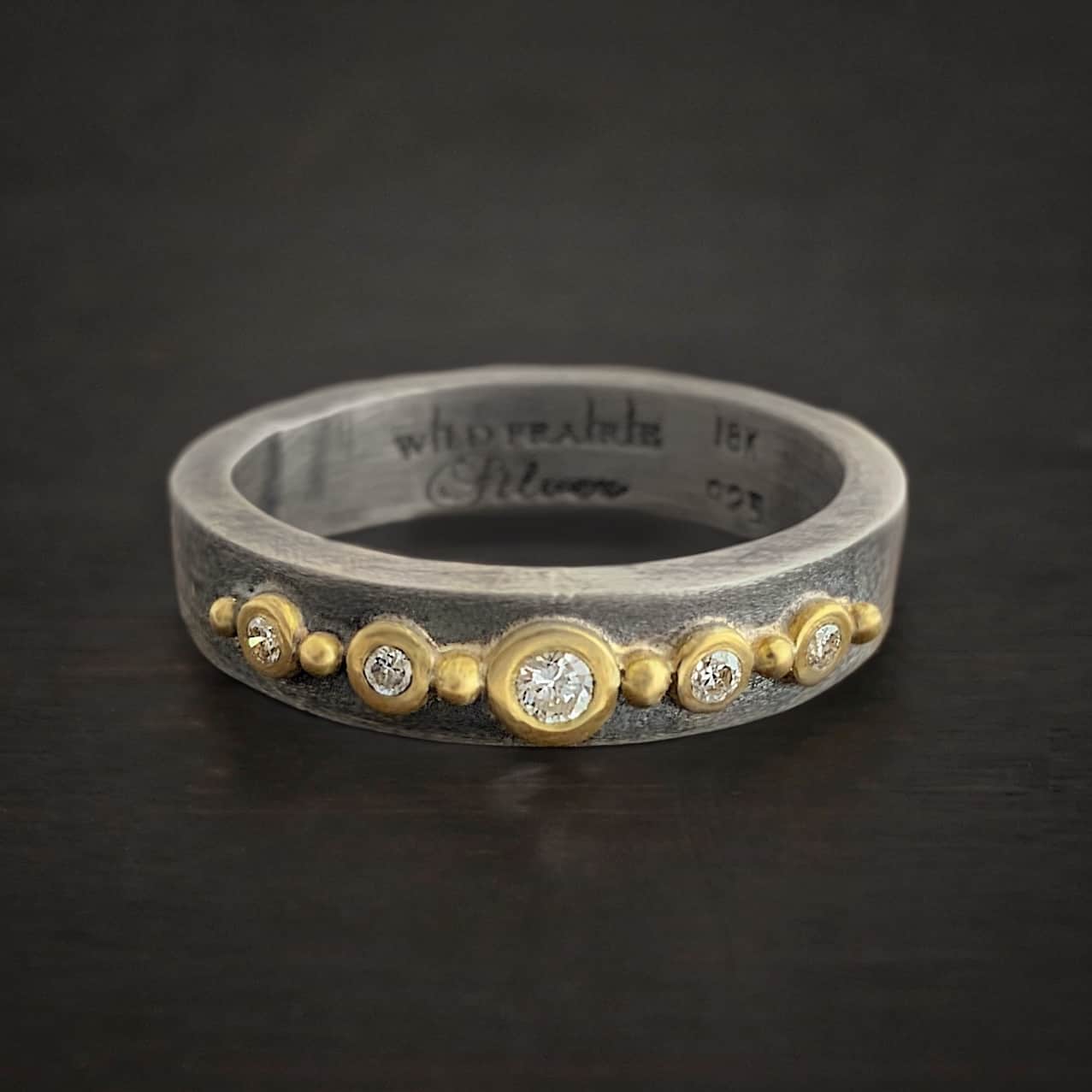 Photo of the "Adoration" Ring. Made out of sterling silver, gold, and diamonds. There are five diamonds that go straight across the center of the ring. Each one is set in gold, and each diamond is separated by a ball of gold. There are six balls of gold in total. The rest of the ring is oxidized and has a textured look to it. The words "Wild Prairie Silver" and "18K" and ".925" are all stamped on the inside of the ring.