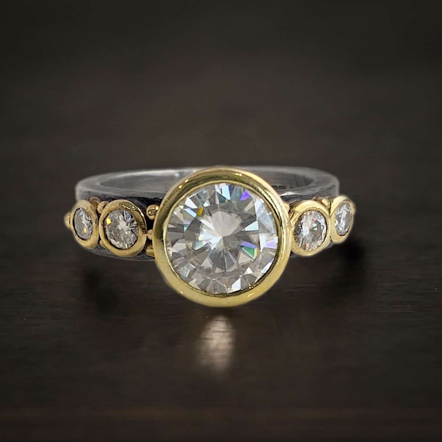 Until Forever Engagement Ring Photo of the "Until Forever" Engagement Ring. There is a large moissanite in the center of the ring. There are four more moissanites that are smaller, two on each side of the center moissanite. There are balls of gold scattered around each moissanite.