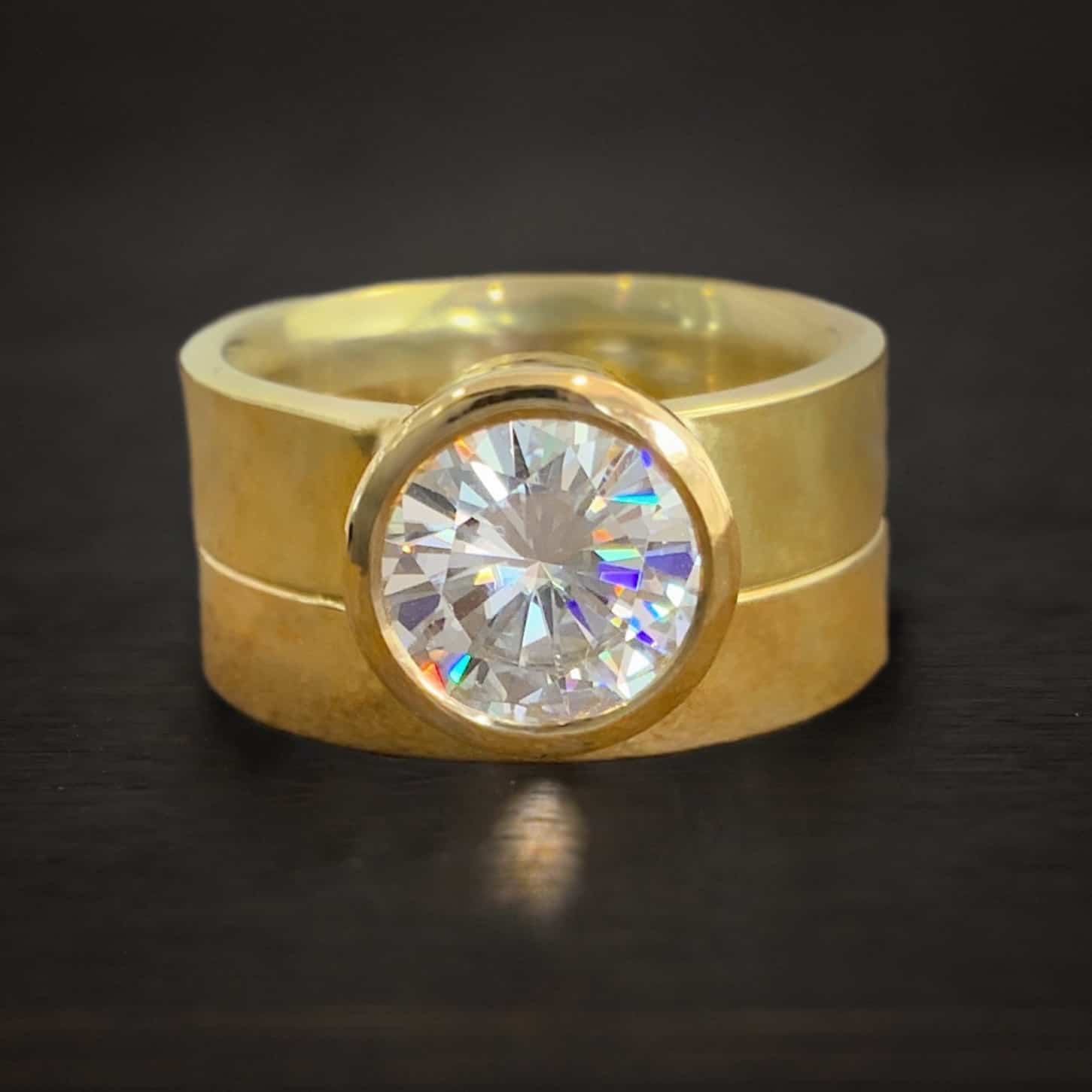Forever After All - All gold Photo of the "Forever After All" Ring. Made out of gold and moissanite. There is a large moissanite in the center of the ring. The band is thick and gold. There is another, thick gold band beneath it with a groove in the center to fit around the stone tightly. Ring is sitting against a dark background.