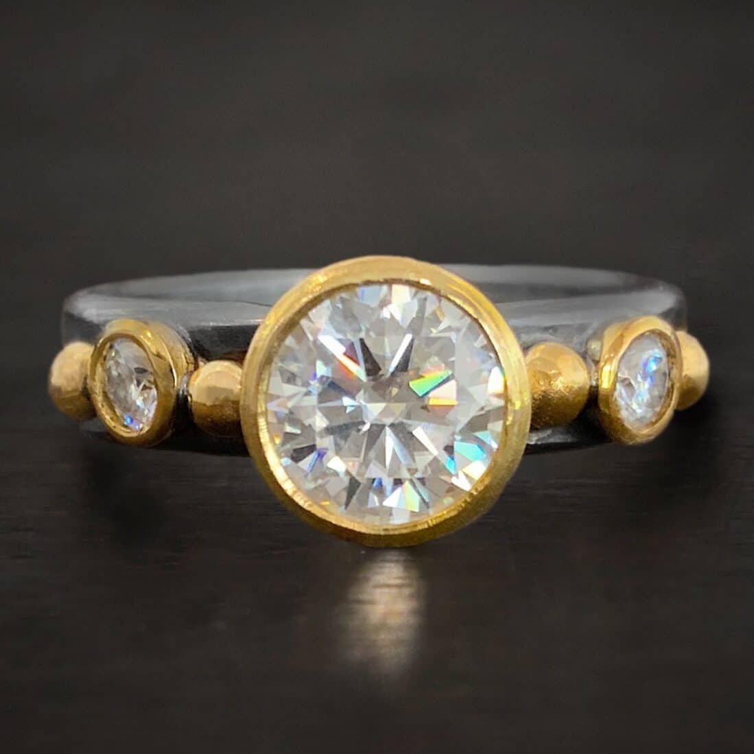 Honeymoon Ring Photo of the "Honeymoon" Ring. Made out of sterling silver, gold, and moissanites. There is a very large moissanite in the center. On each side of it is a ball of gold. After each ball of gold is another, smaller moissanite. After each moissanite is another ball of gold. There are four balls of gold total and three moissanites on a silver band.