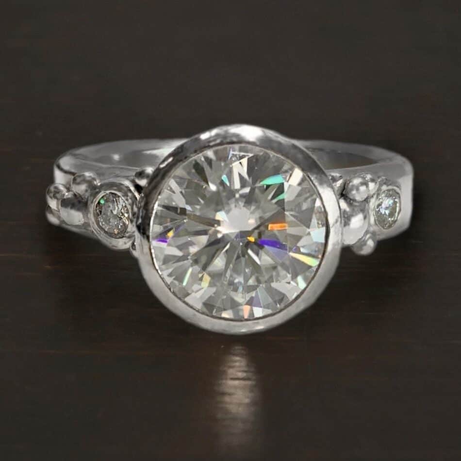 Love Story Engagement Ring Photo of the "Love Story" Ring. Made out of silver and moissanite. There is a large moissanite in the center of the ring that is set in silver. There are two more moissanites, one on each side of the band. The band of the ring has a lot of silver ball detail around it. Both sides of the large moissanite have silver ball detail, as well as around the smaller ones. Ring is sitting against a dark background.