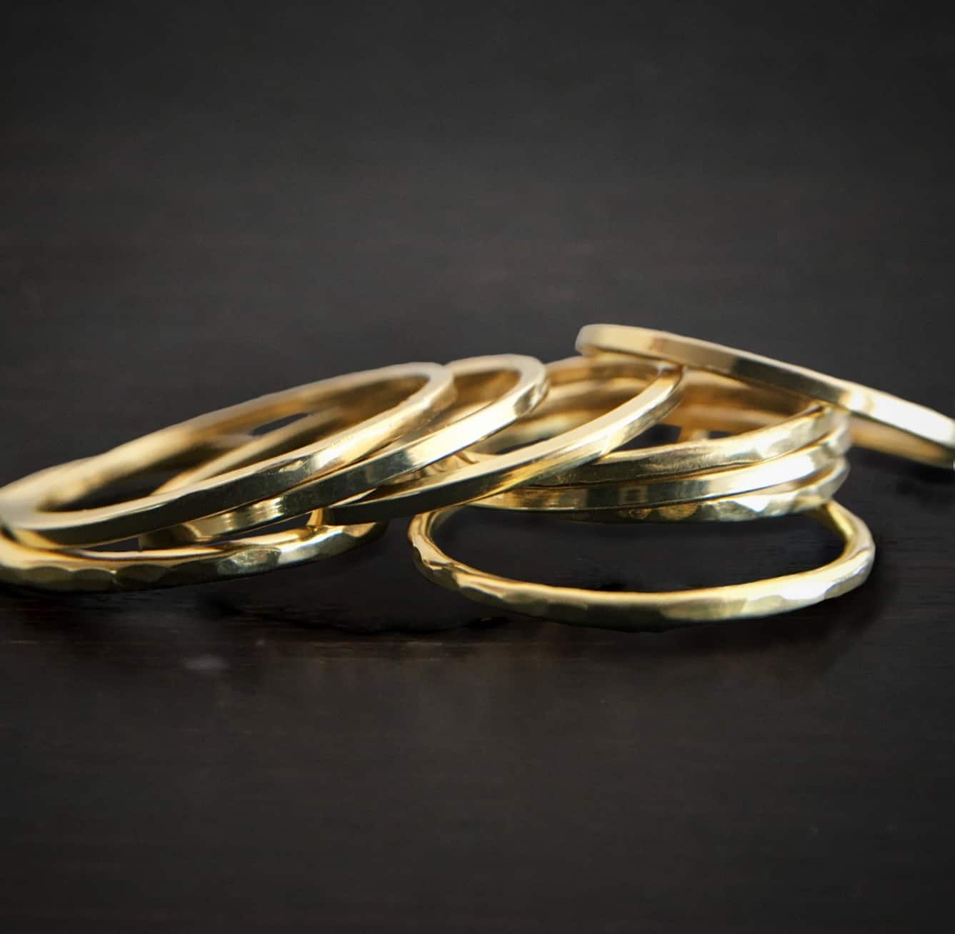 Photo of the "Gabrielle Gold" Stackers. The stackers are thin gold bands that are hammered. There are many that are stacked in a pile. Rings are sitting against a dark background.